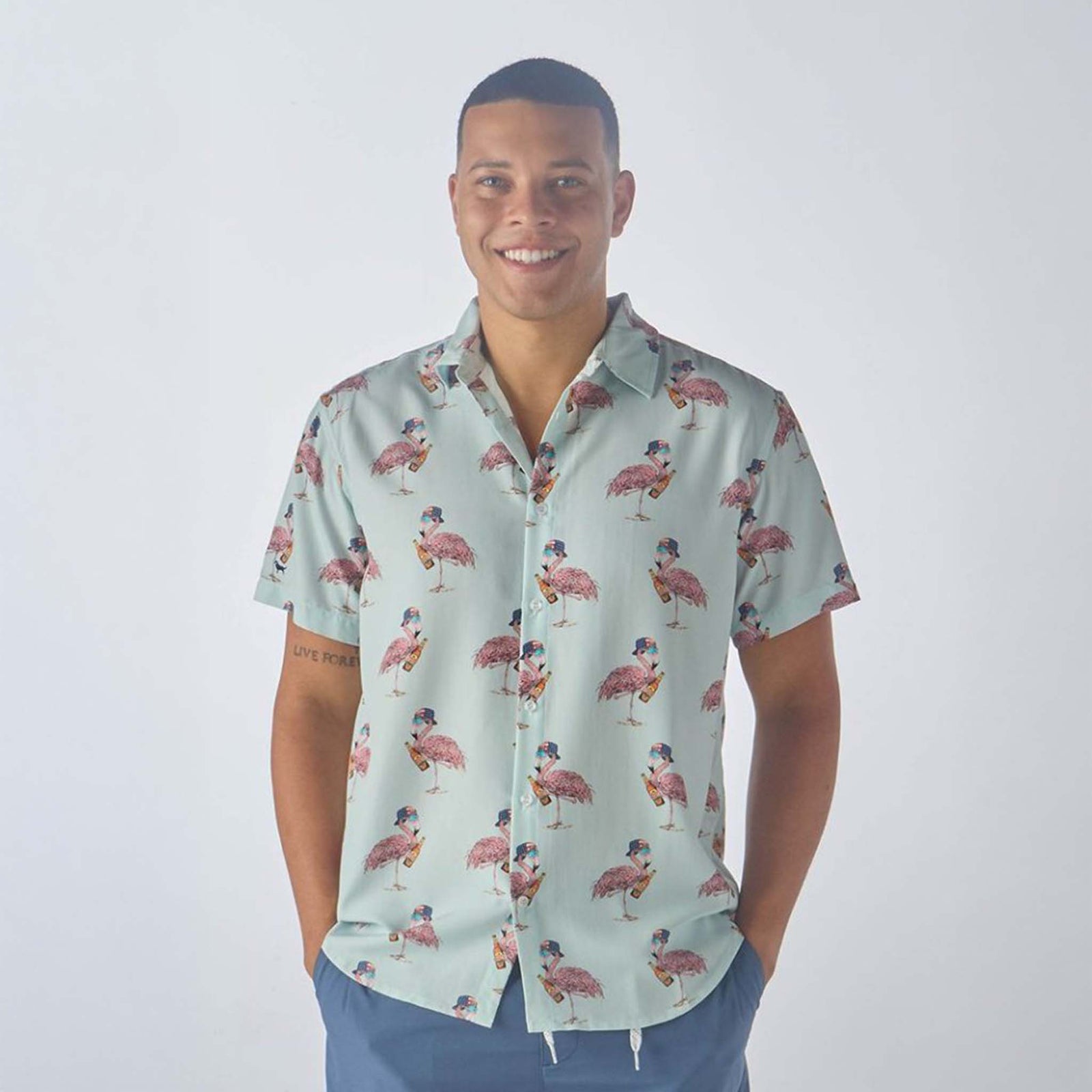 A smiling man wearing a Simply Southern short-sleeve shirt with a light mint green background, patterned with pink flamingos wearing sunglasses and holding beverages.