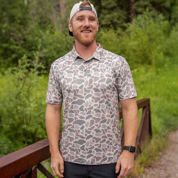 Man wearing Burlebo's Classic Deer Camo Performance Polo outdoors near trees.
