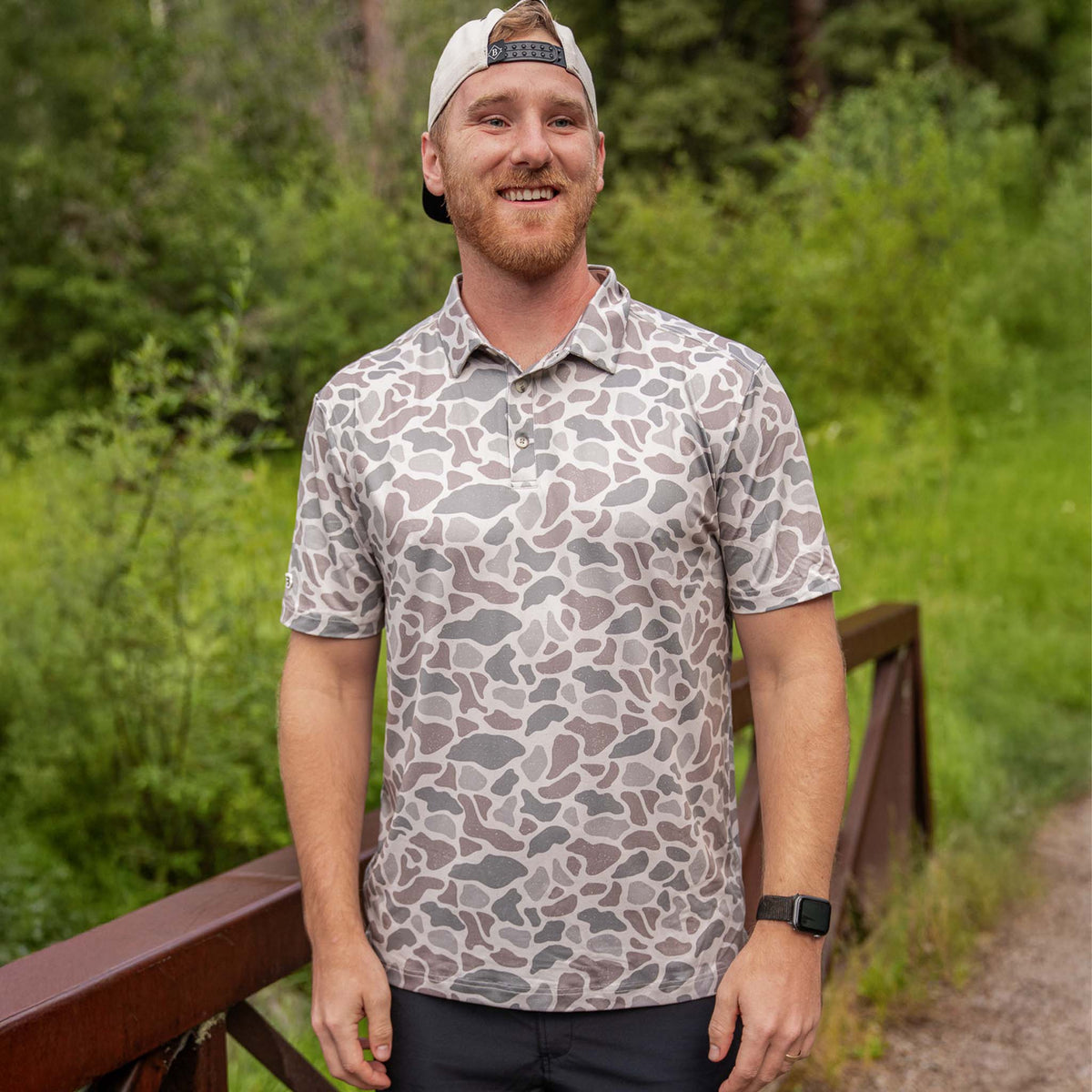 Man wearing Burlebo's Classic Deer Camo Performance Polo outdoors near trees.