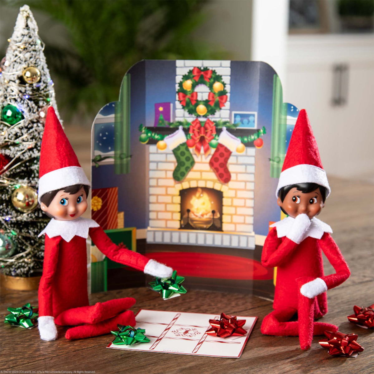 Two Elf on the Shelf dolls with a decorated Christmas scene in the background.