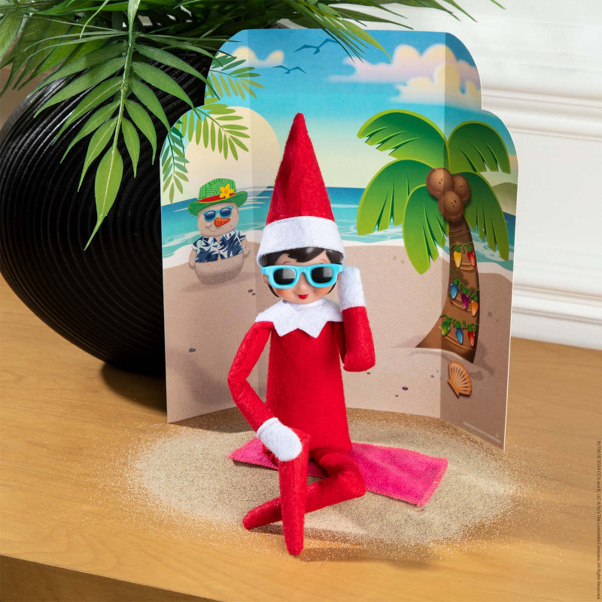 Elf on the Shelf character on a beach with a tropical scene card in the background