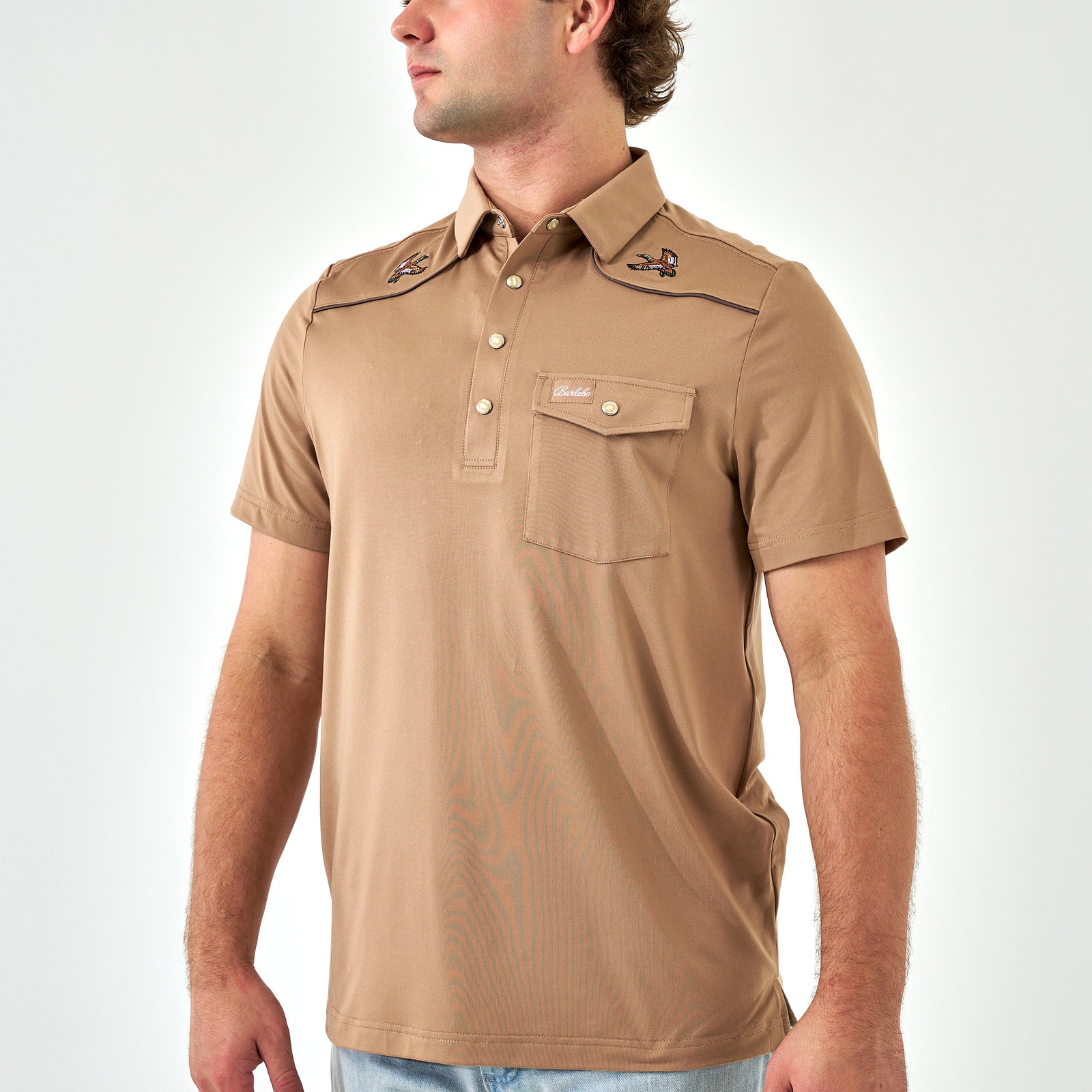 Man wearing a tan short-sleeve polo with brand logos on a white background
