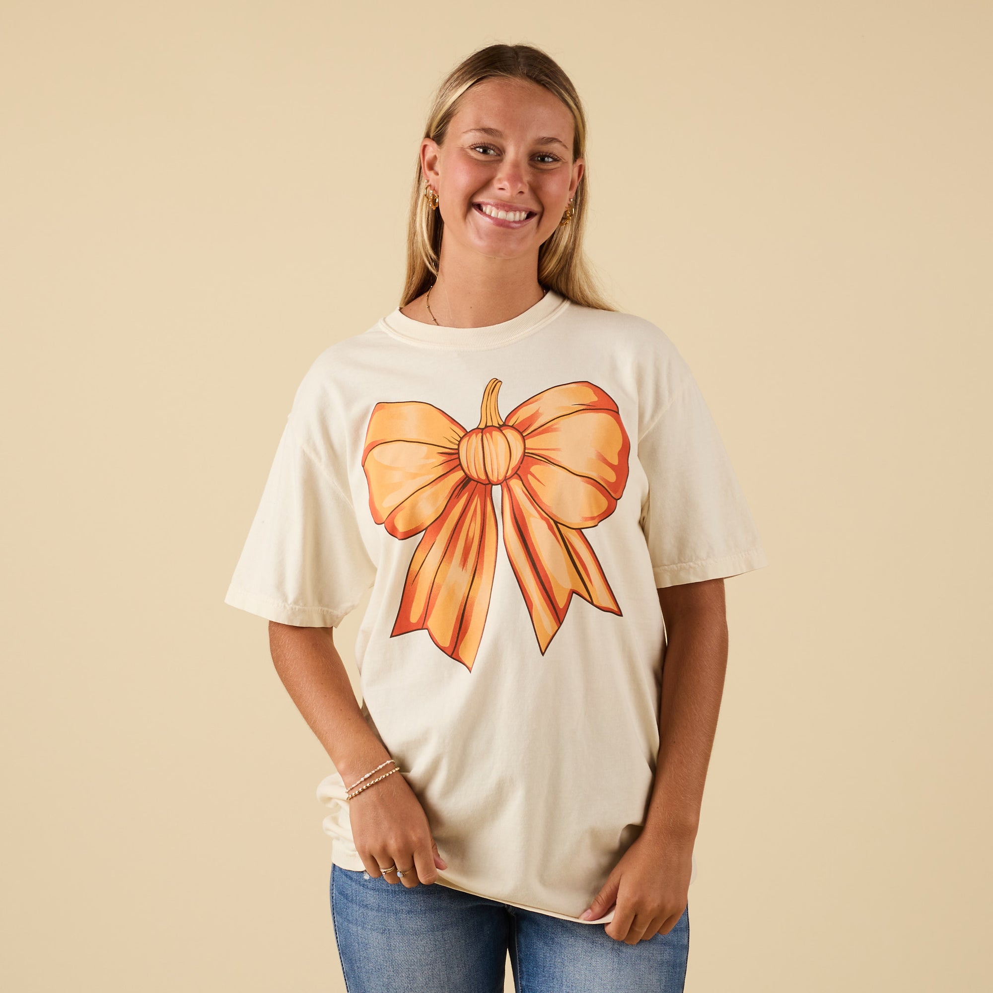Full front view of model wearing ivory t-shirt with oversized pumpkin bow graphic.