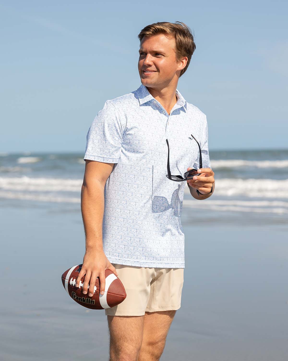 A man standing on the beach wearing a blue polo and khaki shorts while holding a football and sunglasses.