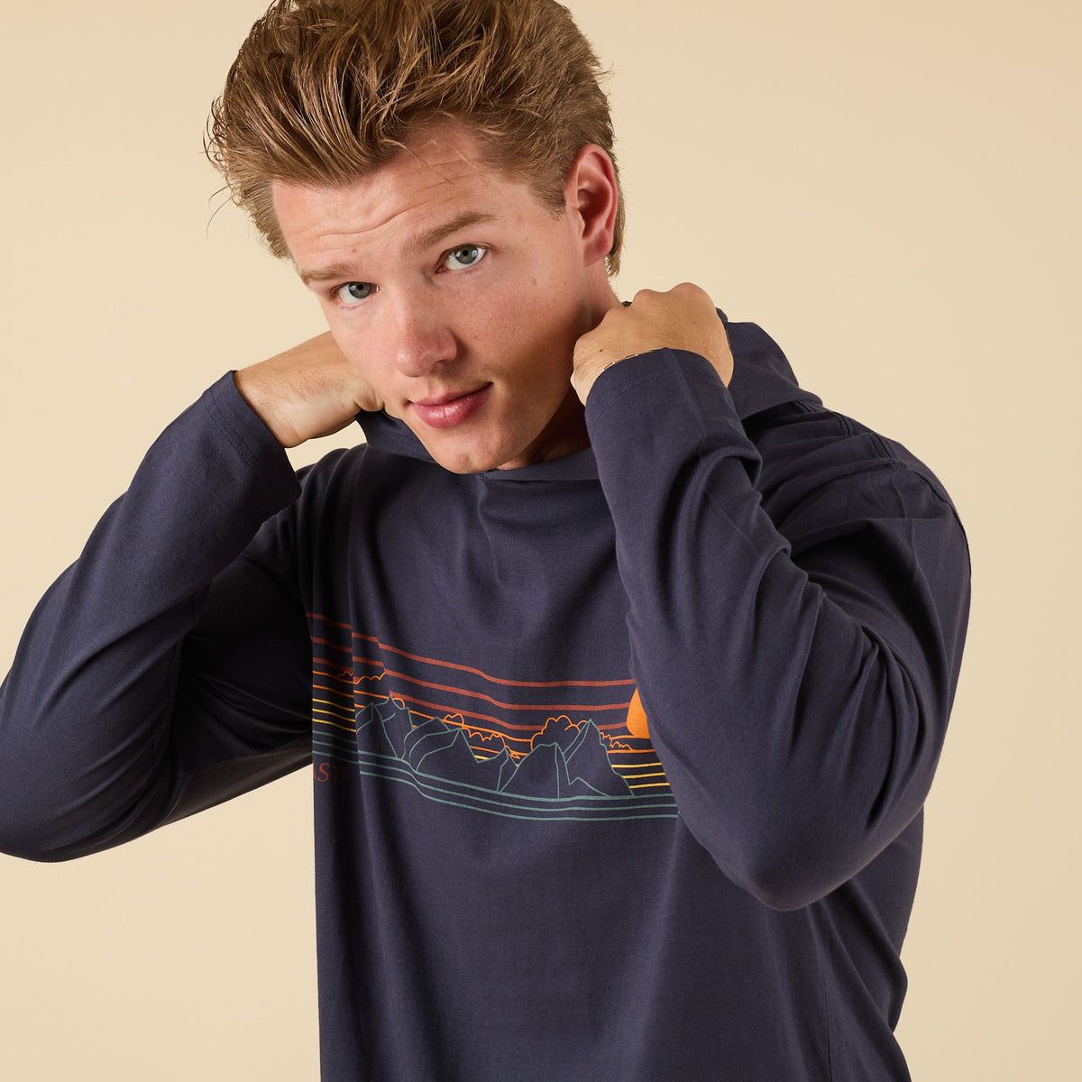 Model adjusting hood while wearing COAST Mountains Hoodie Tee in navy with graphic detail.