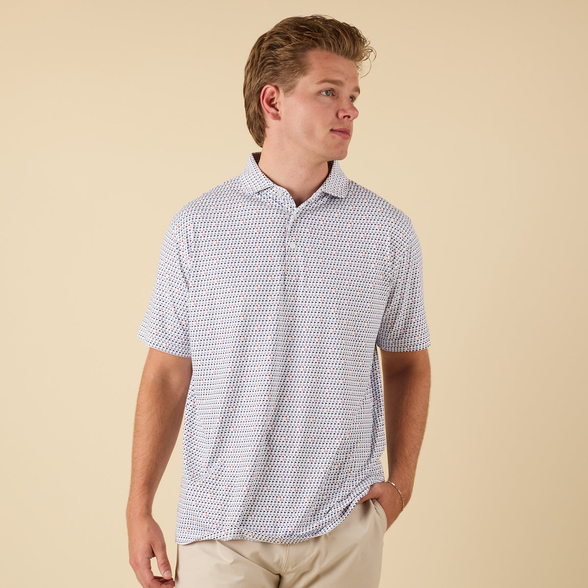 Man wearing a white printed short-sleeve polo shirt with small navy and red golf flag pattern, standing with left hand in pocket facing slightly right.