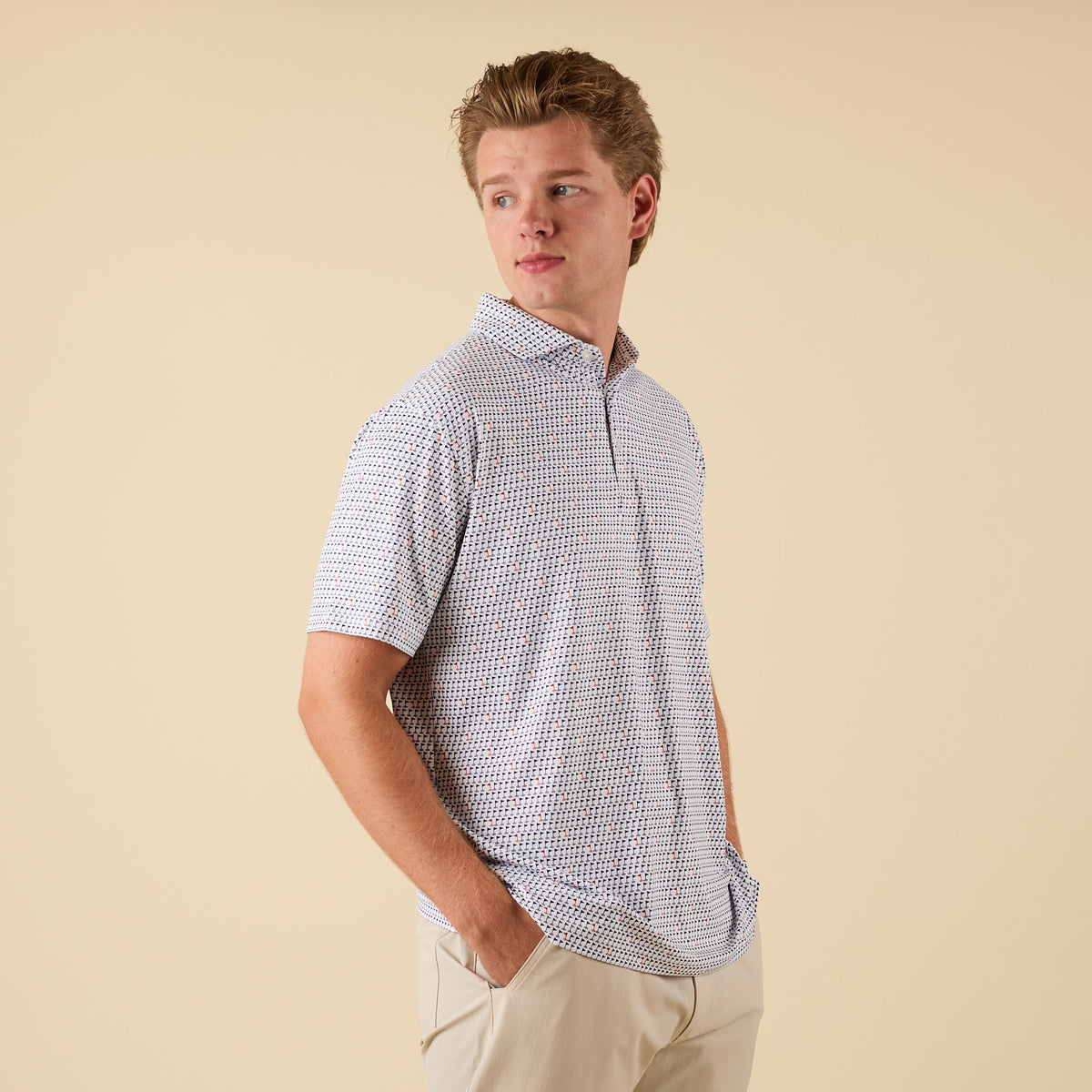 Man in a white short-sleeve printed polo shirt with navy and red golf flag design, standing with hands in pockets looking over left shoulder.