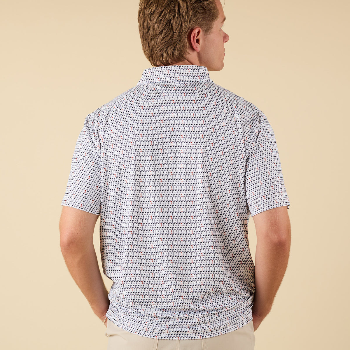 Man wearing a white short-sleeve polo shirt with small navy and red golf flag print, shown from the back with hands in pockets.