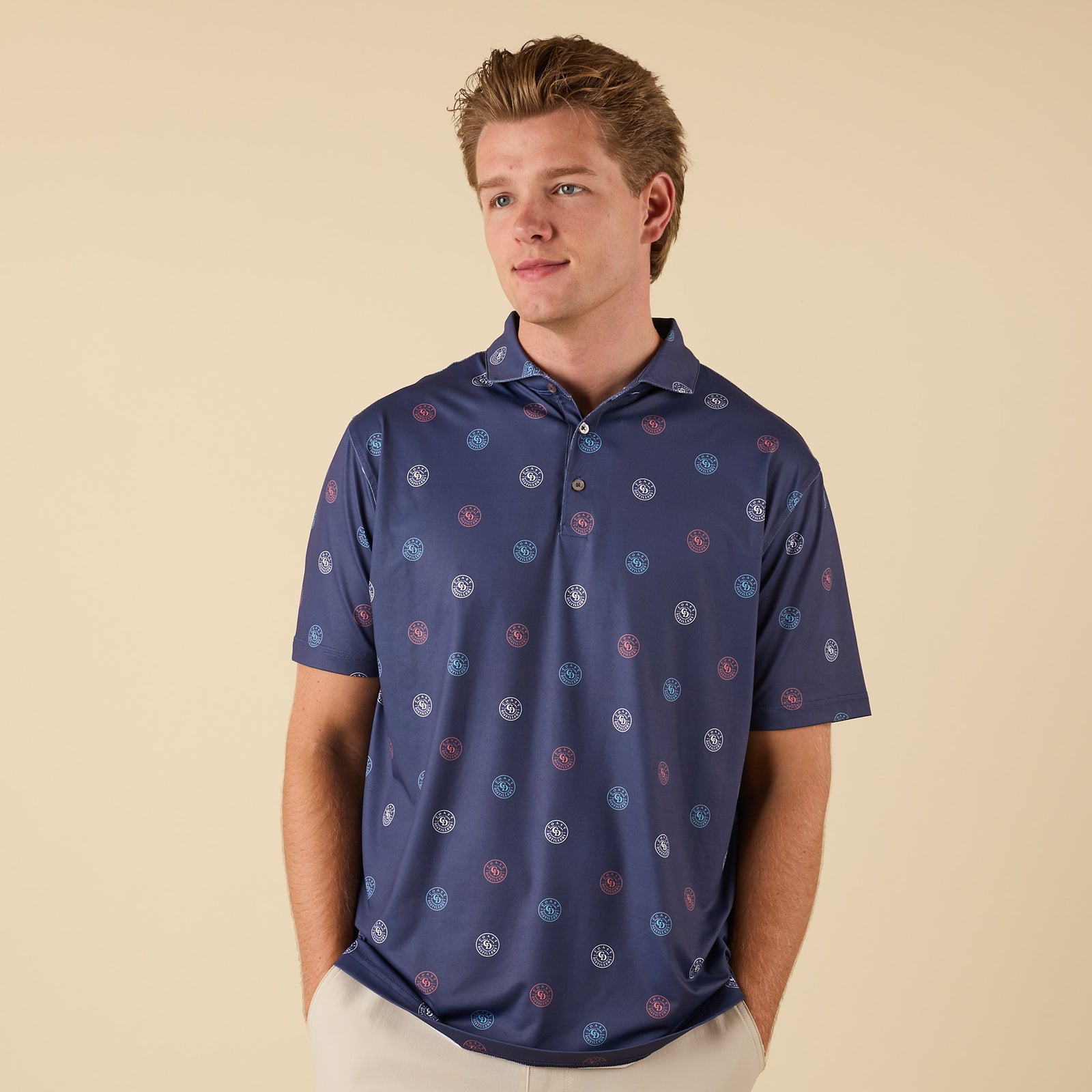 Man wearing a navy polo shirt with Coast circular logo print, standing with hands in pockets facing forward.