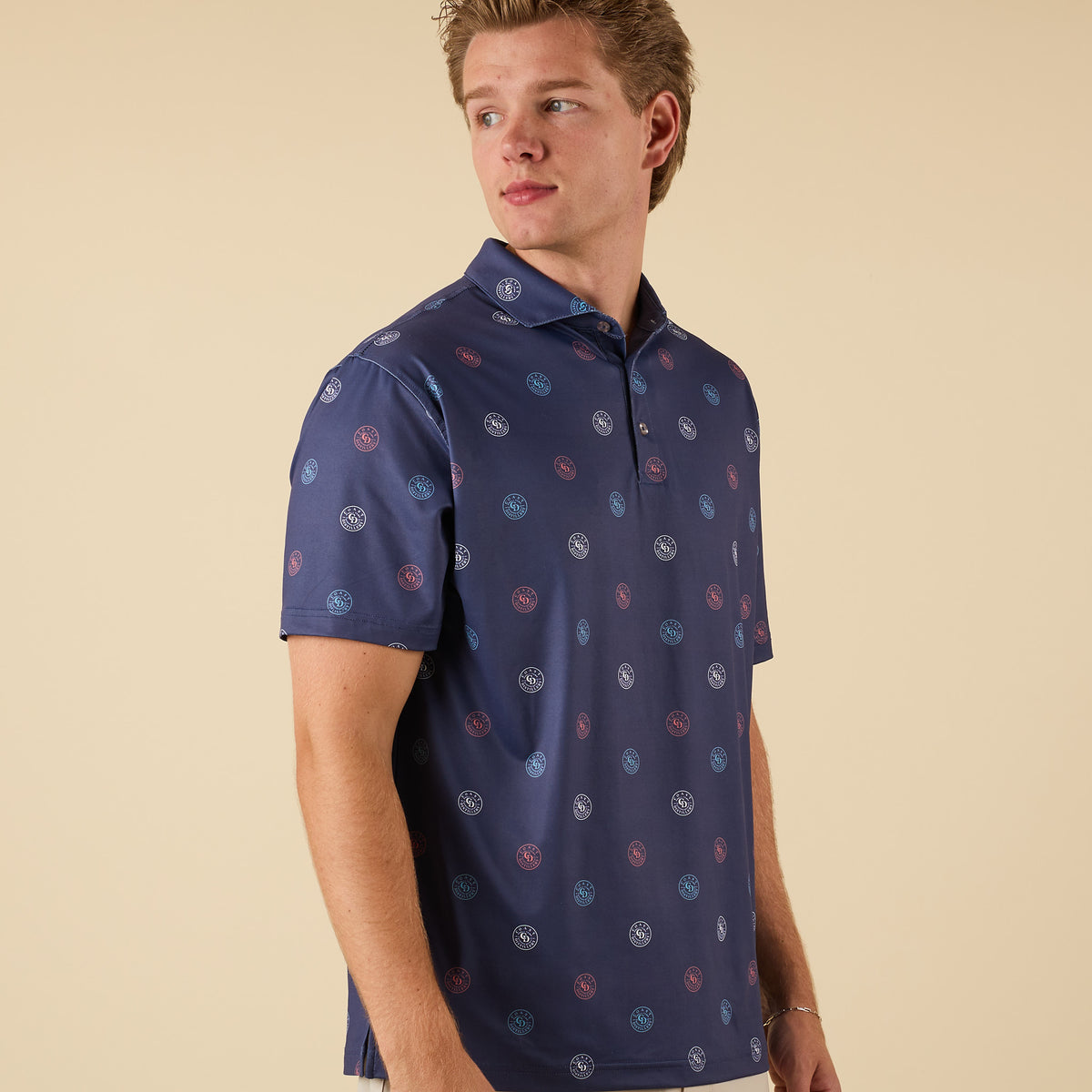 Man wearing a navy polo shirt with all-over Coast circular logo print, standing with arms relaxed looking over left shoulder.