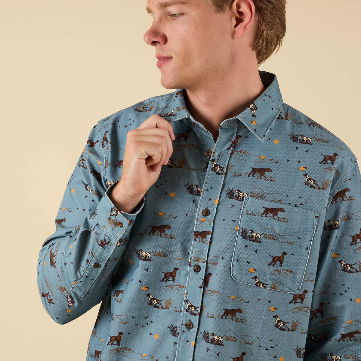 Man adjusting collar in blue hunting scene button-down shirt, side view.
