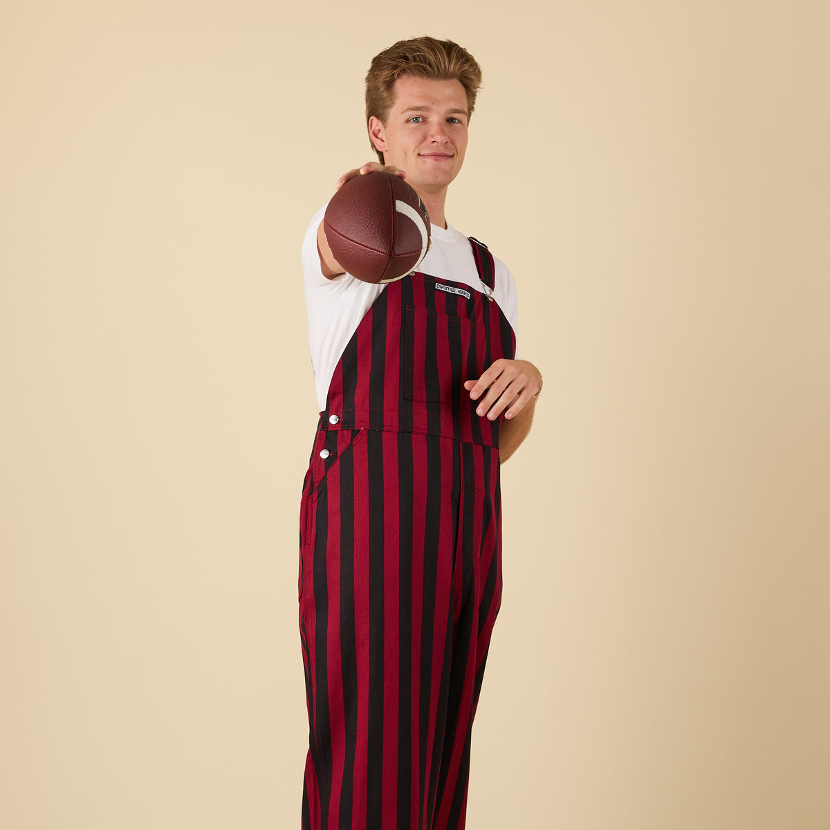 Man posing for a product photo in garnet and black overalls and holding a football up to the camera.