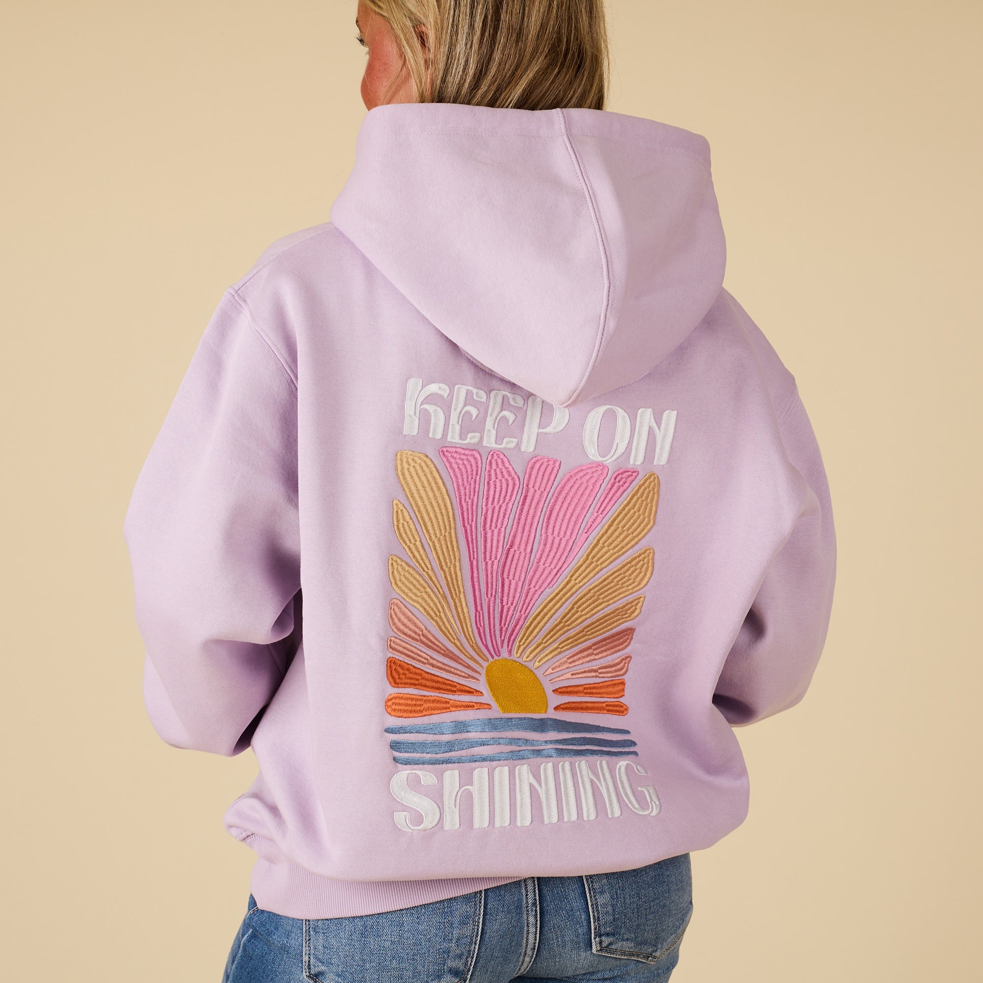 Person wearing a lavender hoodie with a sun graphic and 'Keep On Shining' text on the back, against a beige background.