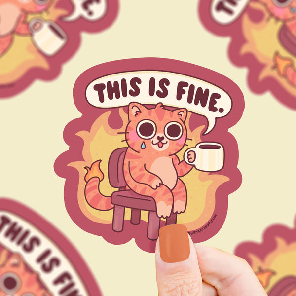 Sticker with a cat holding a coffee cup and 'This is Fine' text, held by a hand.