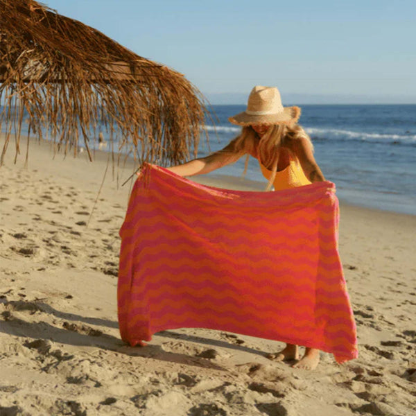 Woman on a beach holding a Pink and orange wavy patterned Sand Cloud towel with white tassels 
