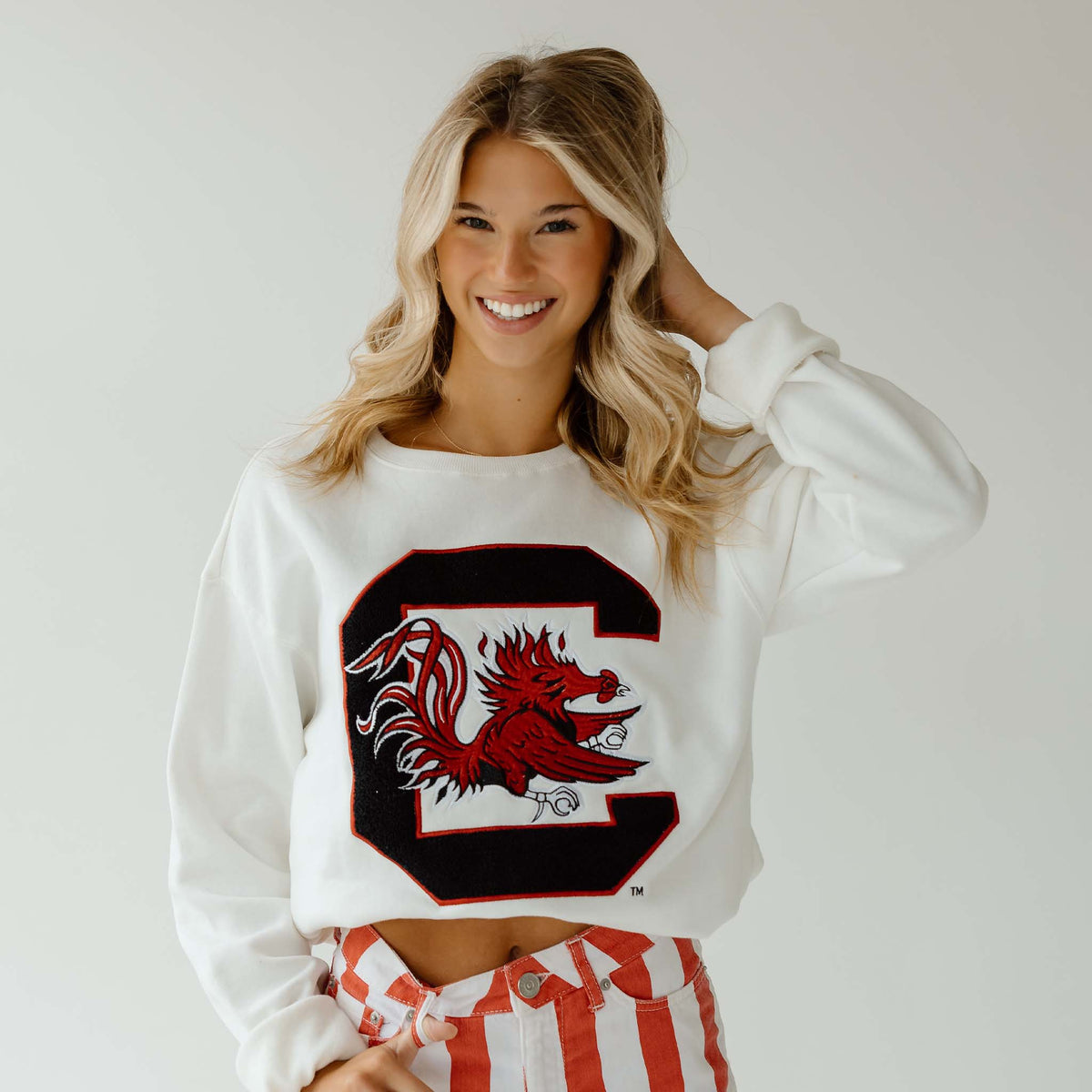 Woman wearing white cropped crewneck sweatshirt with oversized chenille patch University of South Carolina Gamecock logo.