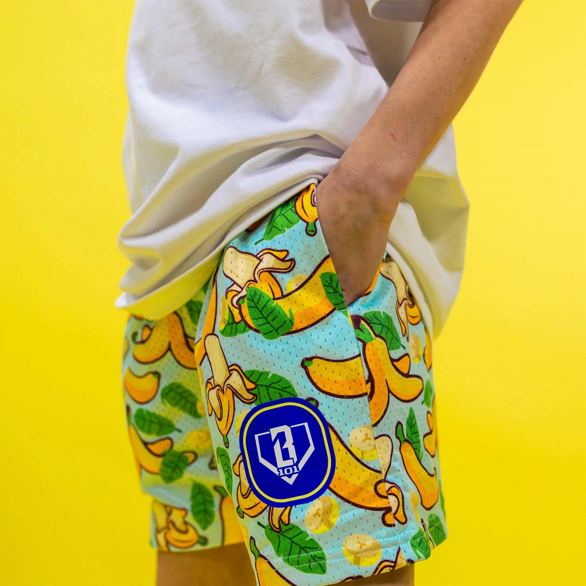 side image of boy wearing of Baseball Lifestyle 101 Youth Shorts tropical banana pattern with his hand in pocket.