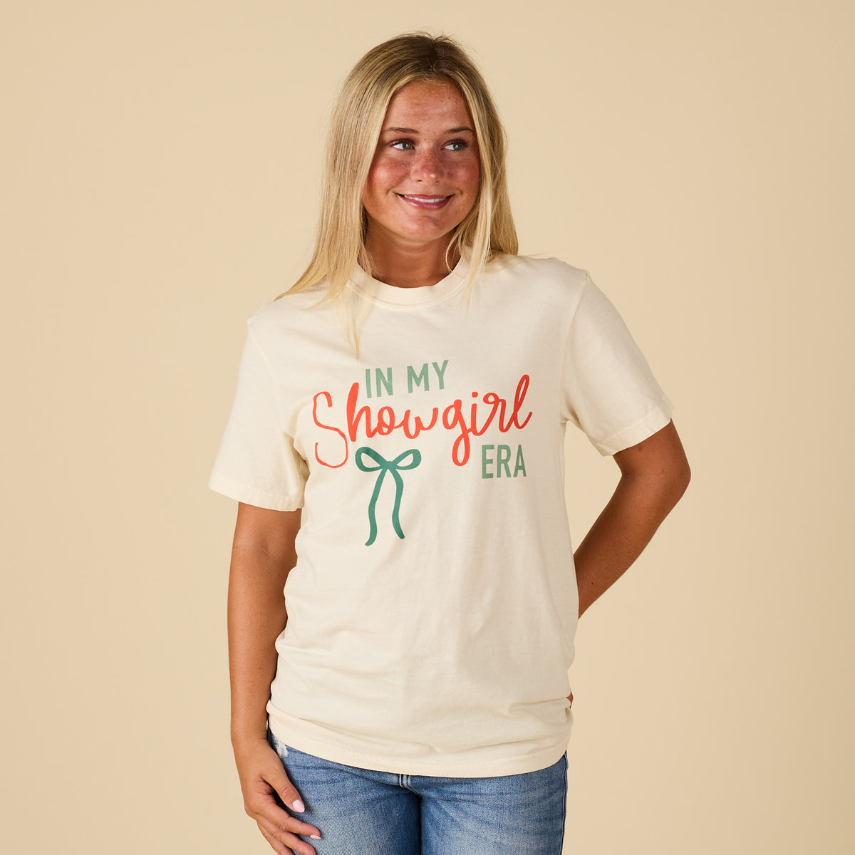 Front view of a woman wearing an ivory short sleeve t-shirt with the text “In My Showgirl Era” in red and green lettering and a green bow graphic, styled with jeans.