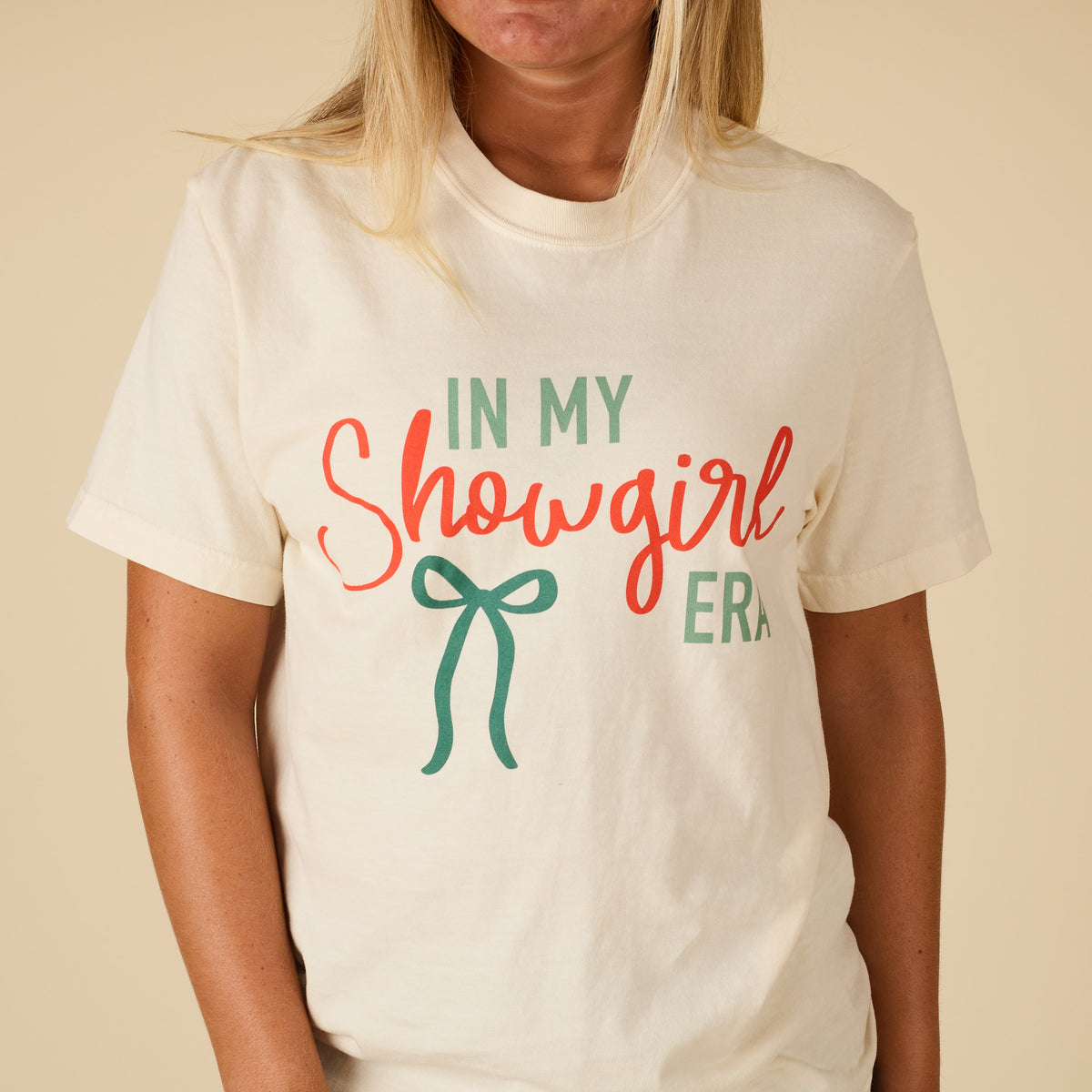 Close-up of the ivory t-shirt front design featuring the phrase “In My Showgirl Era” in bold lettering with a green bow graphic.