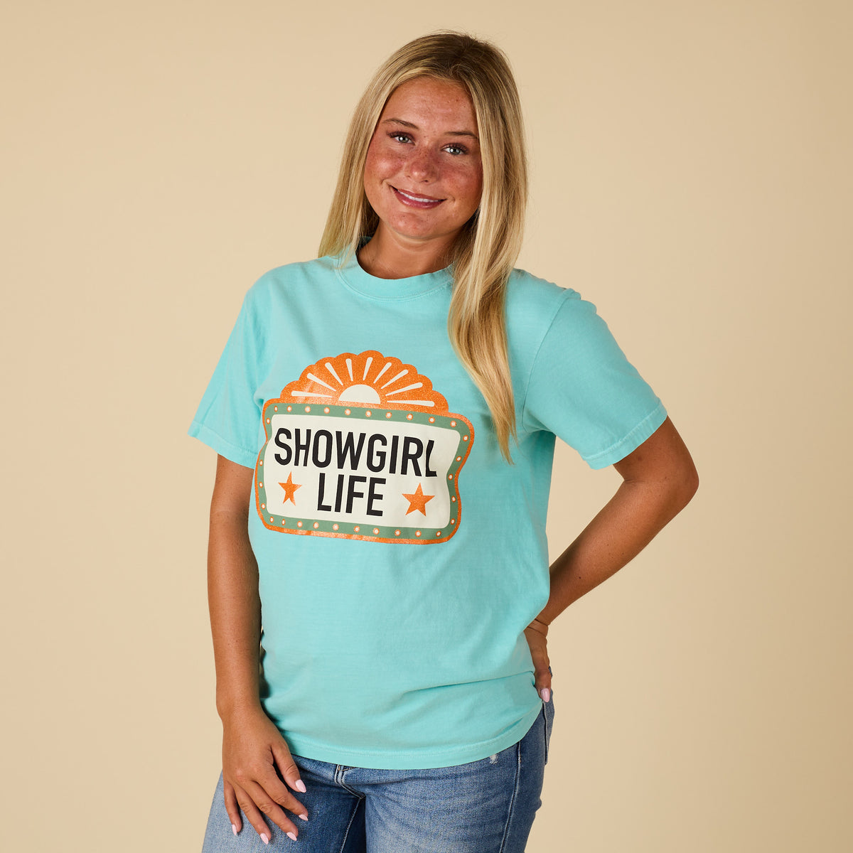 Full front view of a woman wearing the chalky mint “Showgirl Life” graphic t-shirt styled with jeans.
