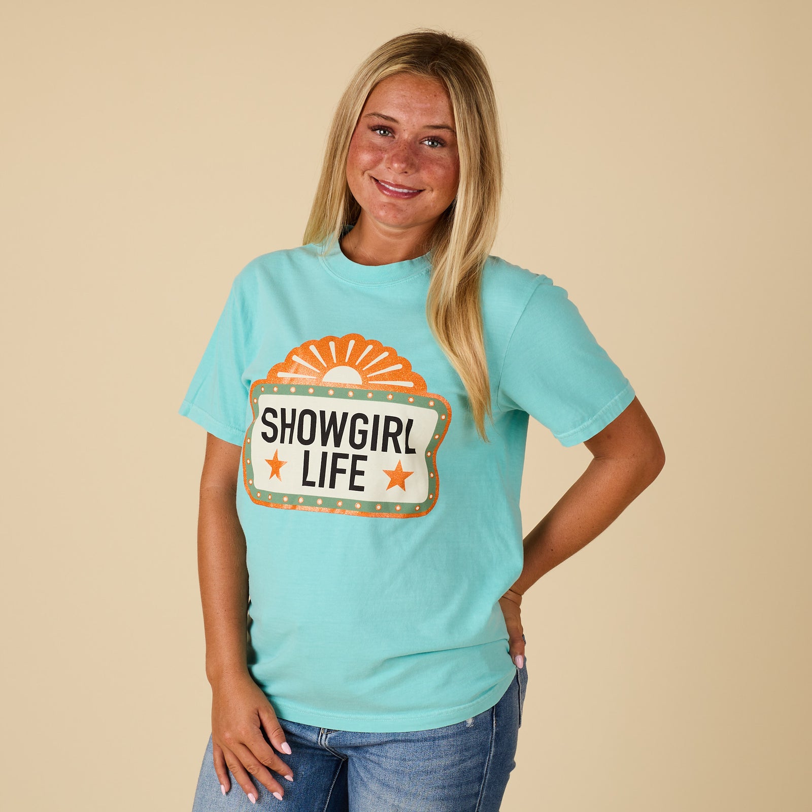 Full front view of a woman wearing the chalky mint “Showgirl Life” graphic t-shirt styled with jeans.