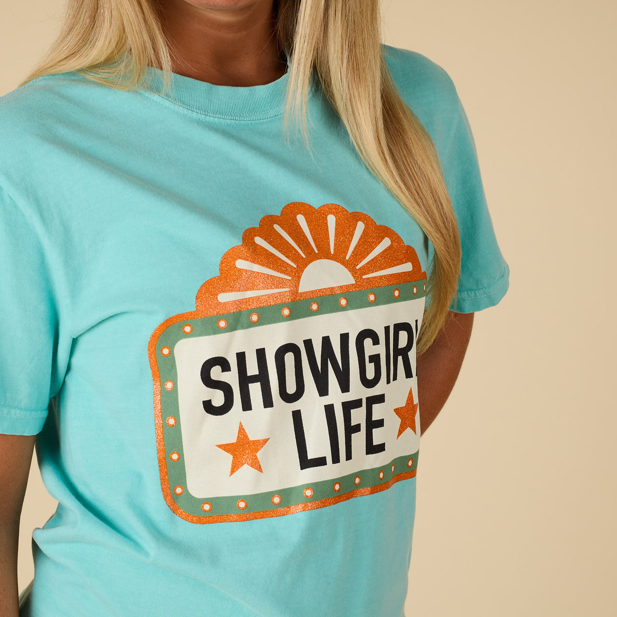Front view of a women’s chalky mint short sleeve t-shirt with a large retro-style sign graphic reading “Showgirl Life” in black text with orange stars and glitter accents.