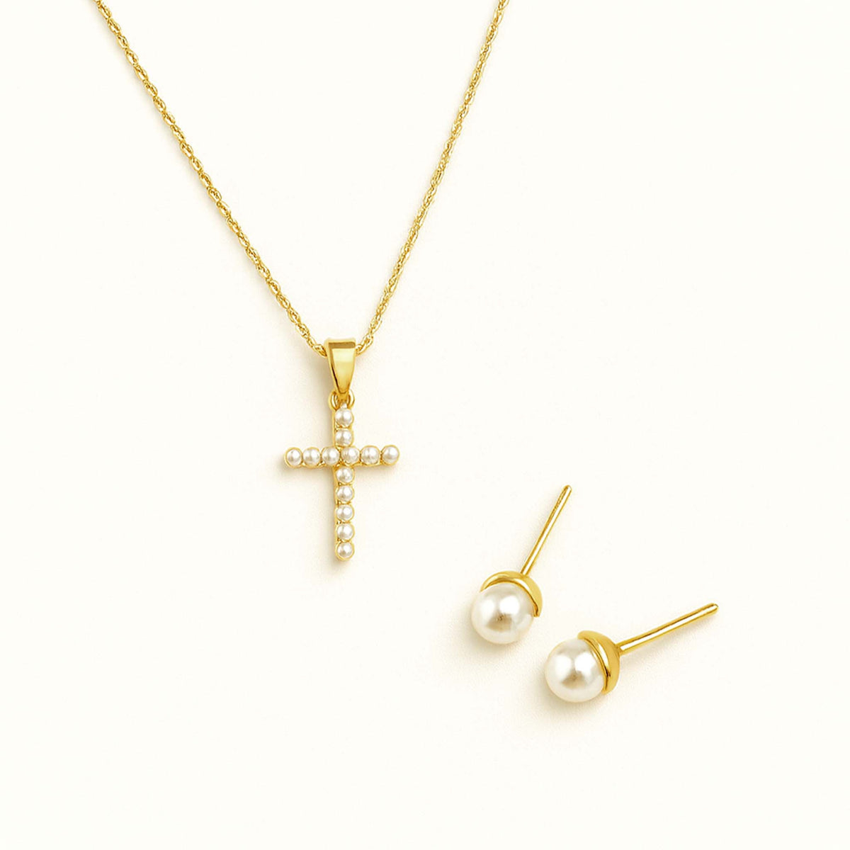 Gold chain with a cubic zirconia cross pendant shown beside matching pearl stud earrings on a cream background.
