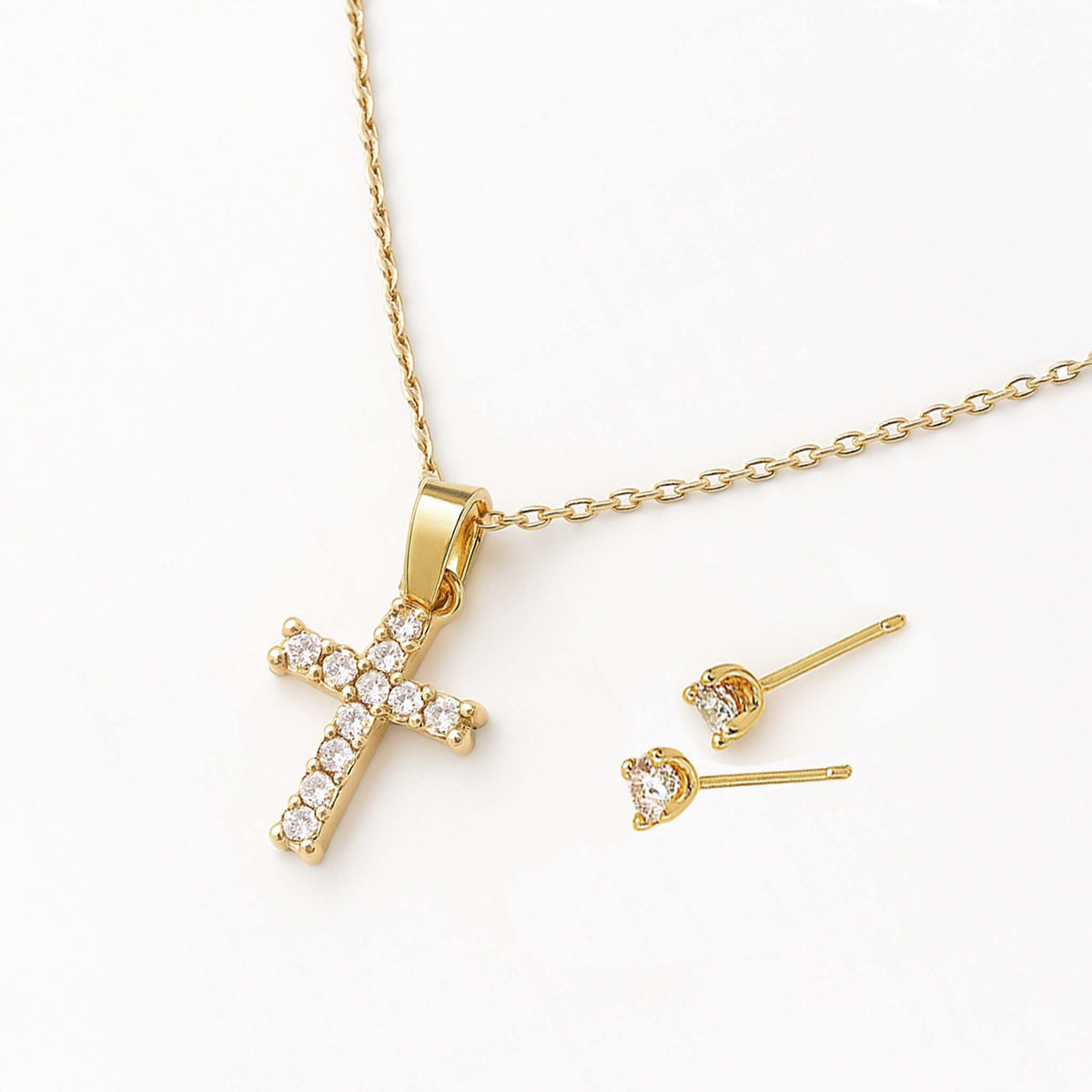 Close-up of a gold cubic zirconia cross necklace shown beside matching CZ stud earrings on a cream background.