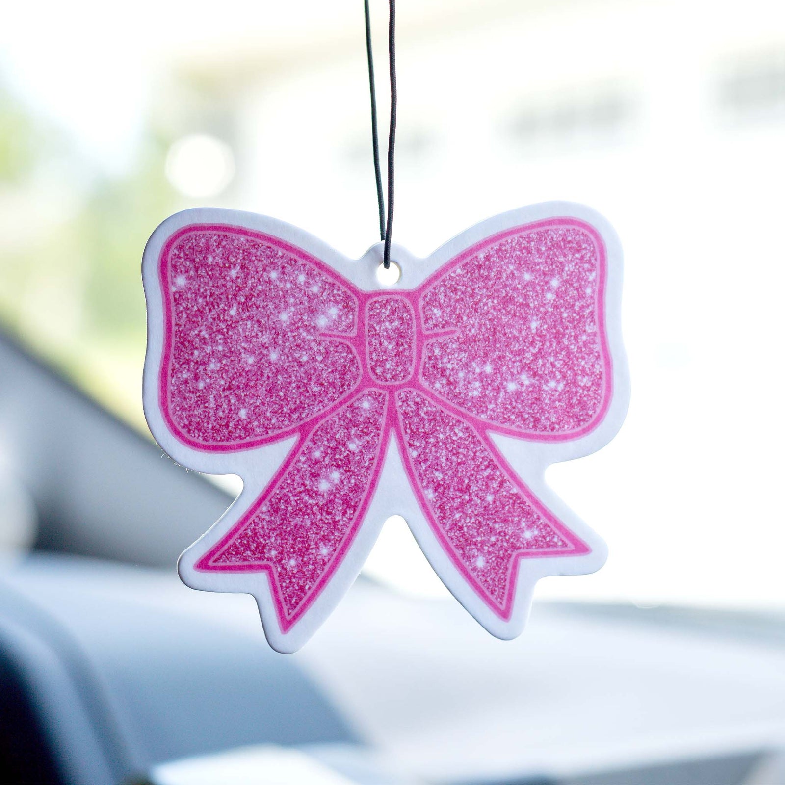 Pink glitter bow-shaped air freshener hanging from a car mirror, photographed with a bright background.