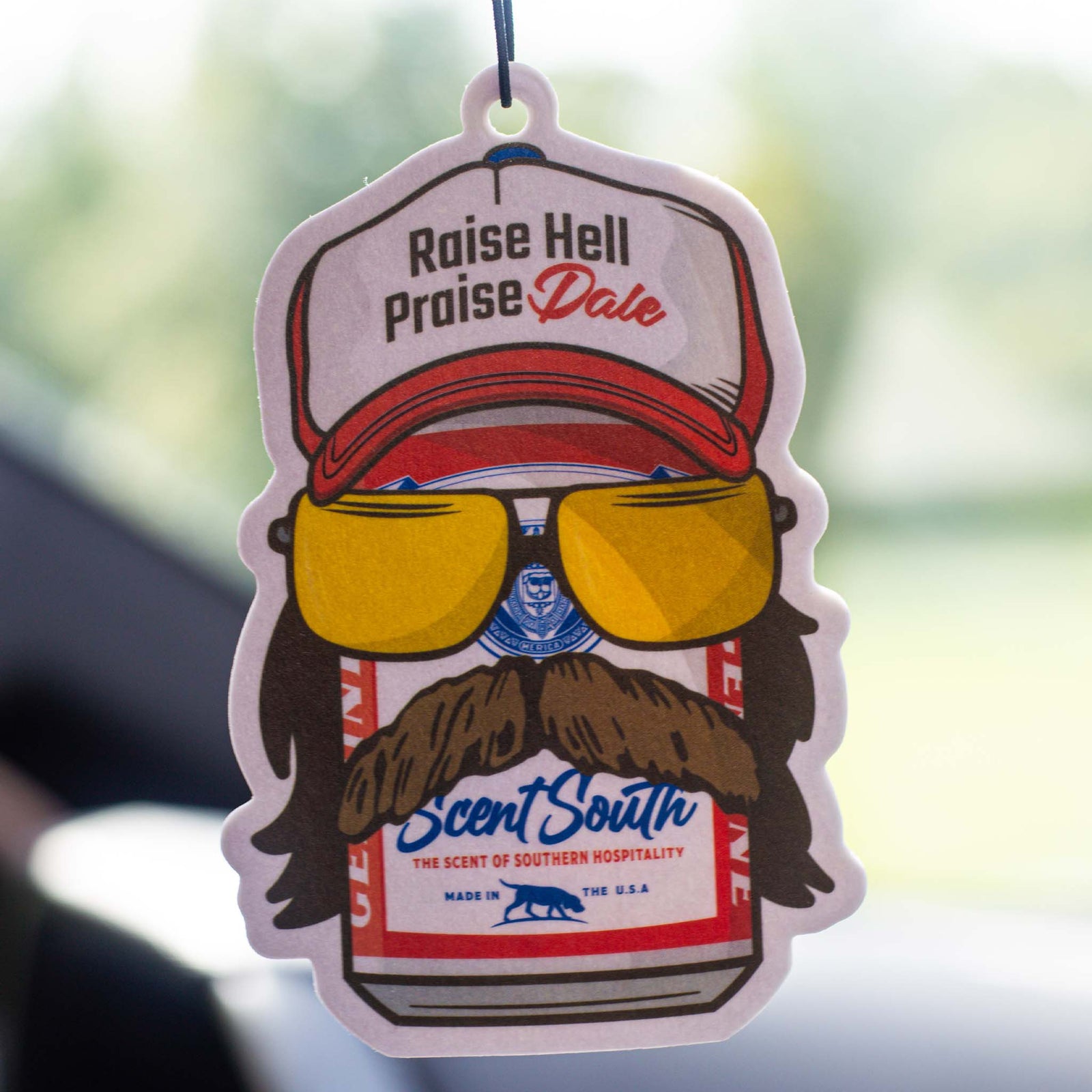 Scent South Praise Dale Hanging air freshener in leather scent.
The design features a graphic of a beer can wearing a red and white hat, yellow lens sunglasses, a mullet, and large mustache.  The hat states "raise hell, praise Dale"