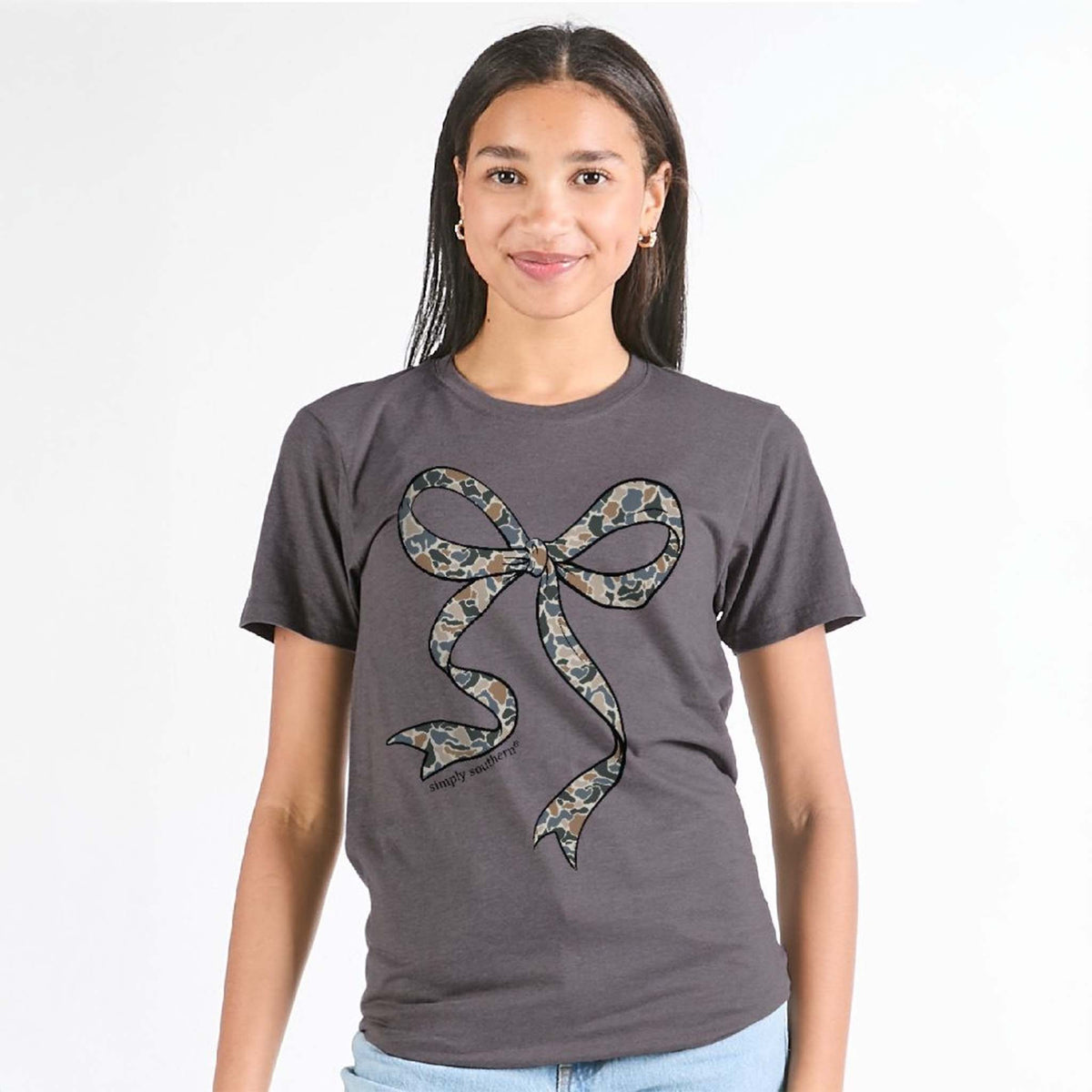 Woman wearing Simply Southern iron heather t-shirt with large camo bow graphic on front.