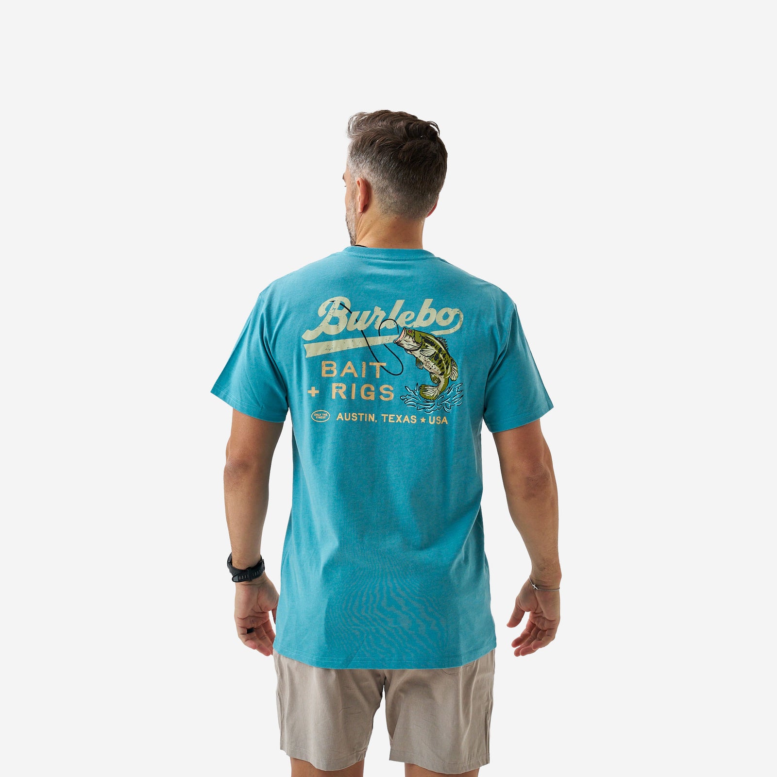Back view of a male model wearing a teal short sleeve graphic t-shirt featuring a “Burlebo Bait + Rigs Austin, Texas USA” fishing design with a largemouth bass illustration.