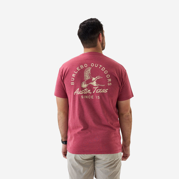 Rear full-body view of a male model wearing a heather red short sleeve Burlebo t-shirt with large duck graphic paired with khaki shorts.