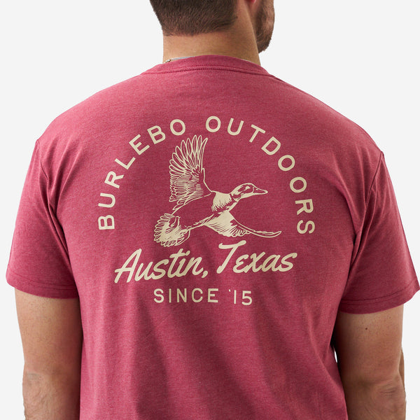 Back view of a male model wearing a heather red short sleeve Burlebo t-shirt featuring a large “Burlebo Outdoors Austin, Texas Since ’15” duck graphic.