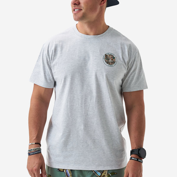 Male model wearing a light gray short sleeve t-shirt with a small circular duck graphic on the chest that reads “Into the Wild Since ’15.”