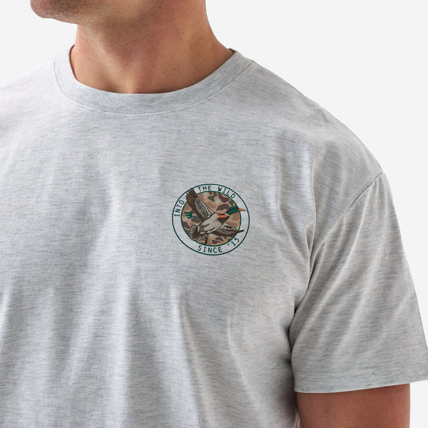 Close-up of a light gray short sleeve t-shirt showing the small chest logo featuring a flying duck and “Into the Wild Since ’15” text.