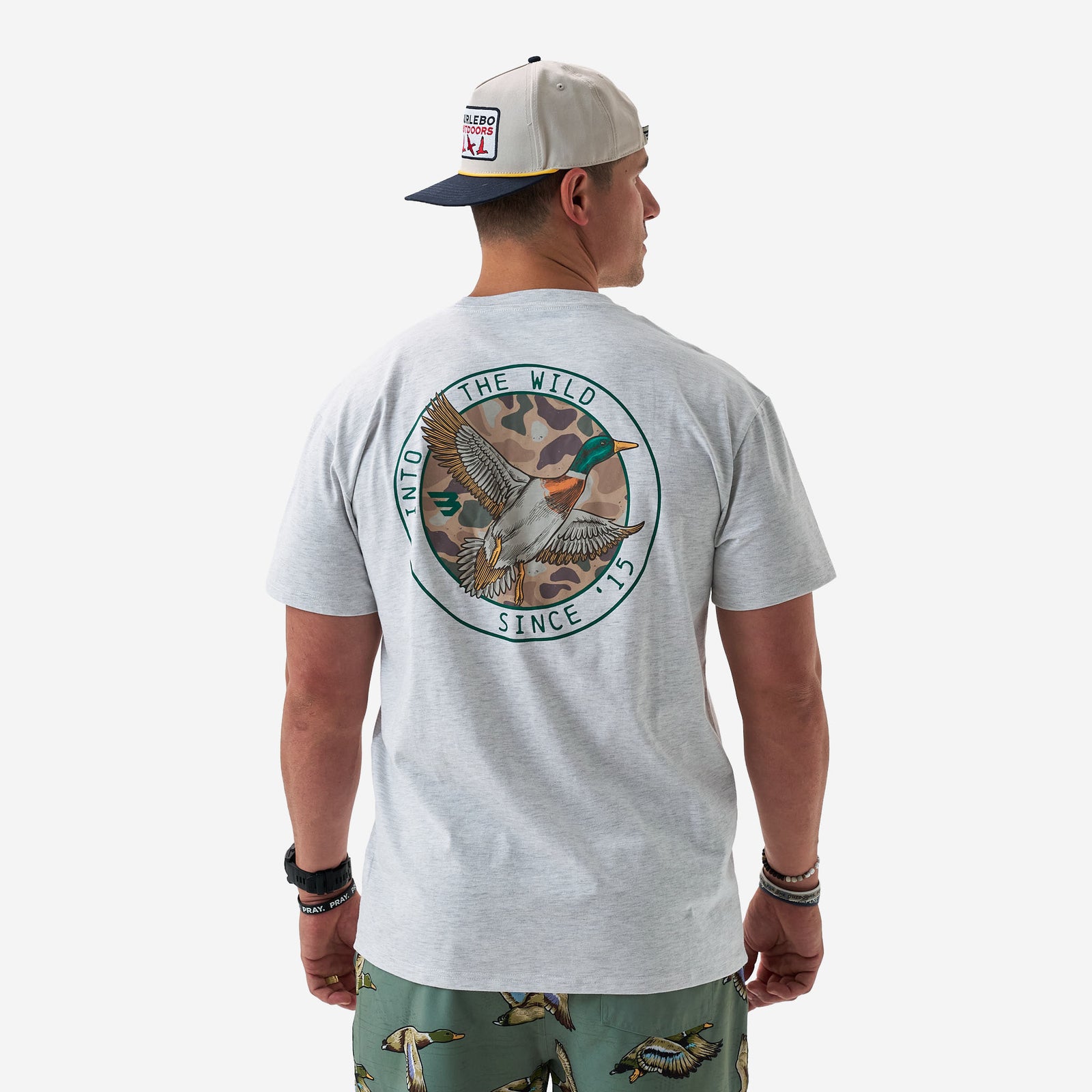 Back view of a male model wearing a light gray short sleeve t-shirt featuring a large circular duck graphic with camouflage background and “Into the Wild Since ’15” text.