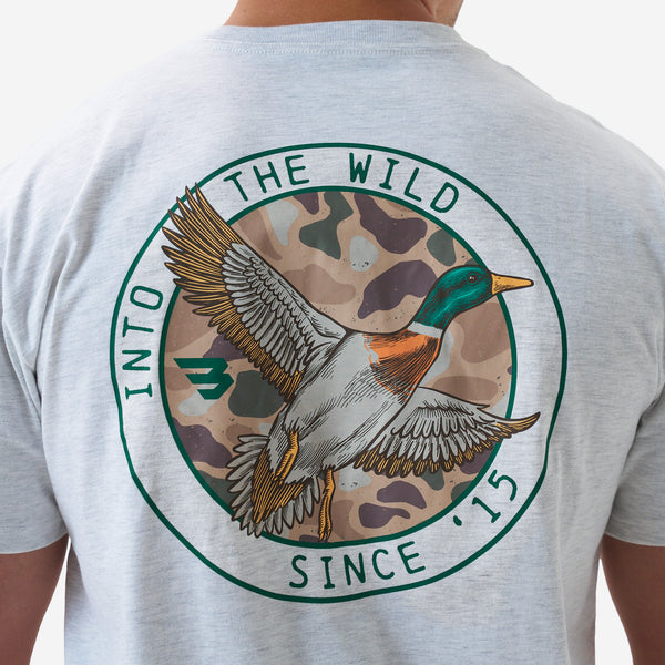 Close-up of the back graphic on a light gray short sleeve t-shirt showing a flying duck illustration inside a circular “Into the Wild Since ’15” design with camouflage background.