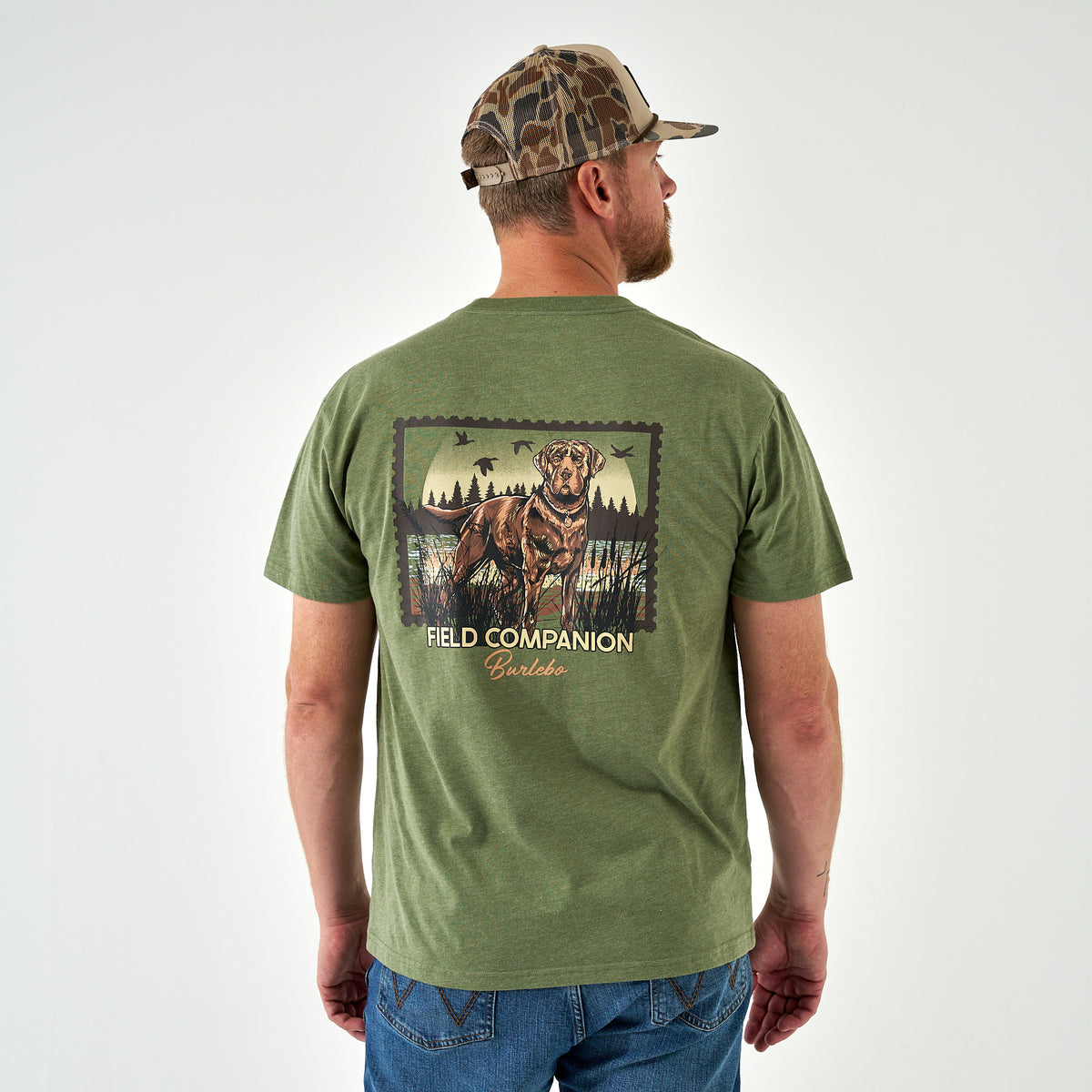 Man wearing an olive green Burlebo Field Companion t-shirt showing the large back graphic of a hunting dog, forest, and flying ducks.vvvvvvv