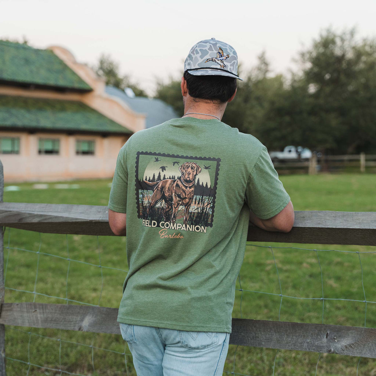 Man leaning on a fence wearing an olive green Burlebo Field Companion t-shirt with a large back graphic of a hunting dog in a field scene.