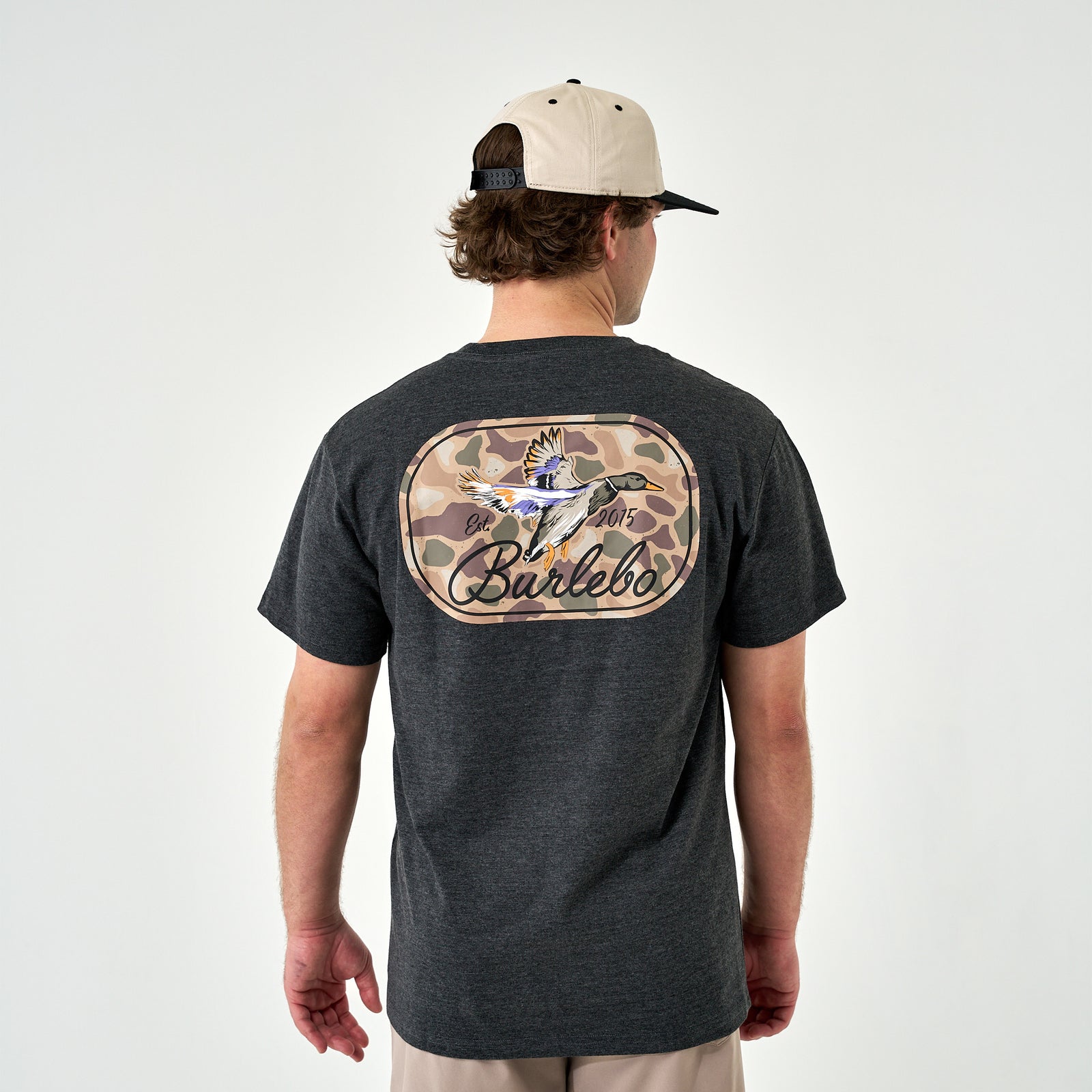 Back view of the dark heather gray t-shirt on a man wearing a beige and black cap, featuring a large camo duck graphic and “Burlebo” text.