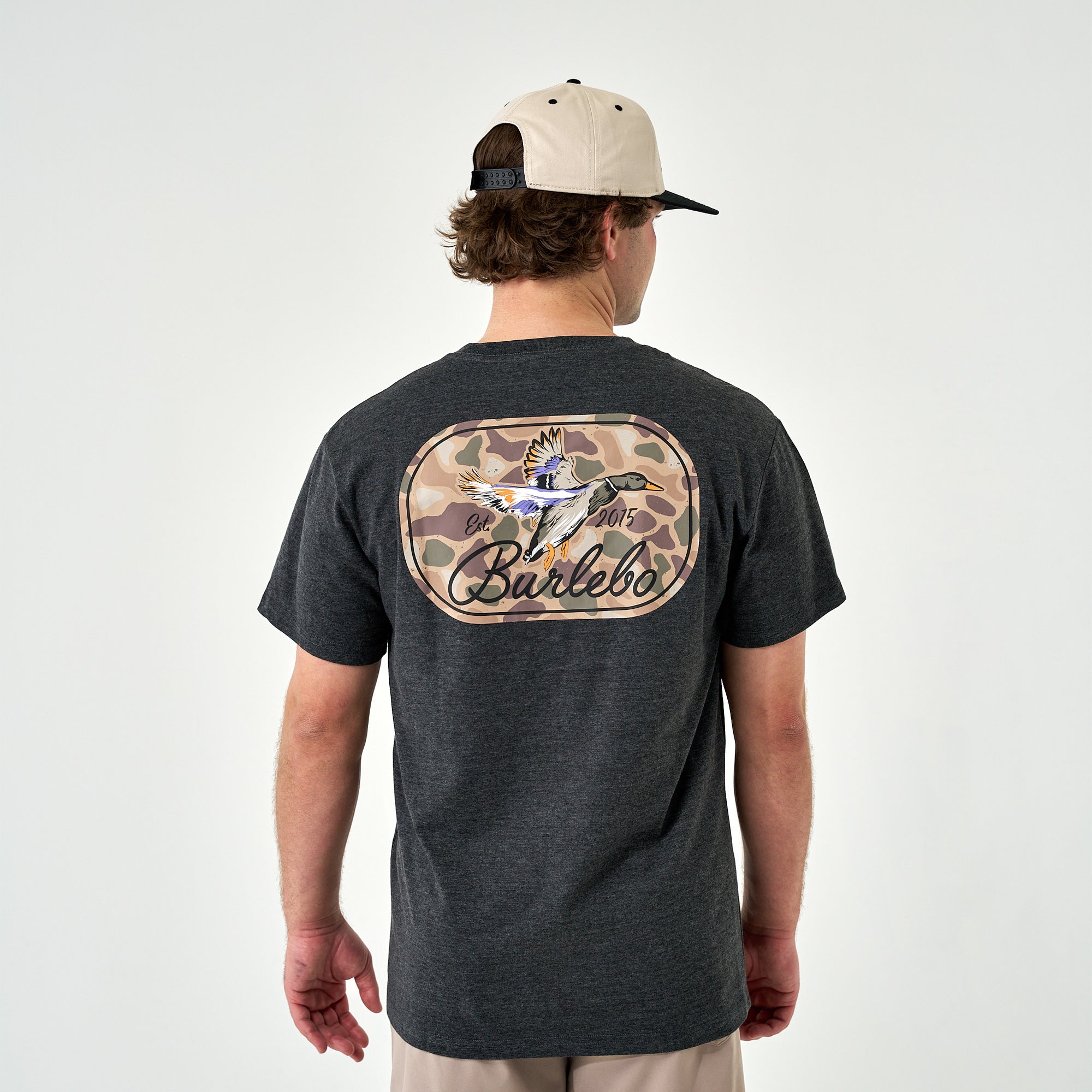 Back view of the dark heather gray t-shirt on a man wearing a beige and black cap, featuring a large camo duck graphic and “Burlebo” text.