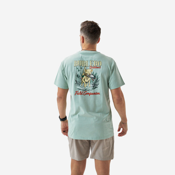 Back view of a male model wearing a sage green short sleeve Burlebo t-shirt featuring a “Burlebo Outdoors Field Companion” graphic with a retriever holding a duck in a marsh scene.