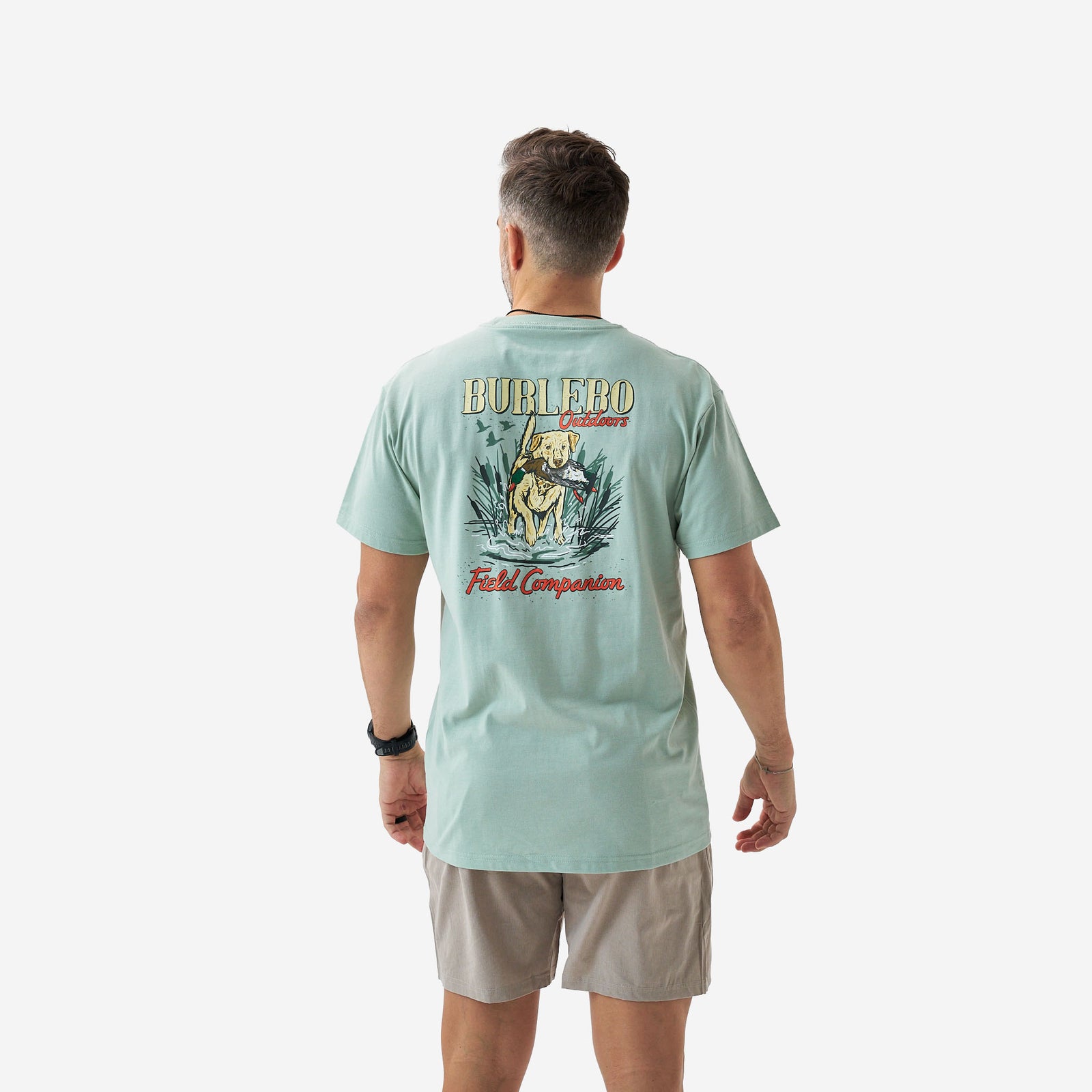 Back view of a male model wearing a sage green short sleeve Burlebo t-shirt featuring a “Burlebo Outdoors Field Companion” graphic with a retriever holding a duck in a marsh scene.