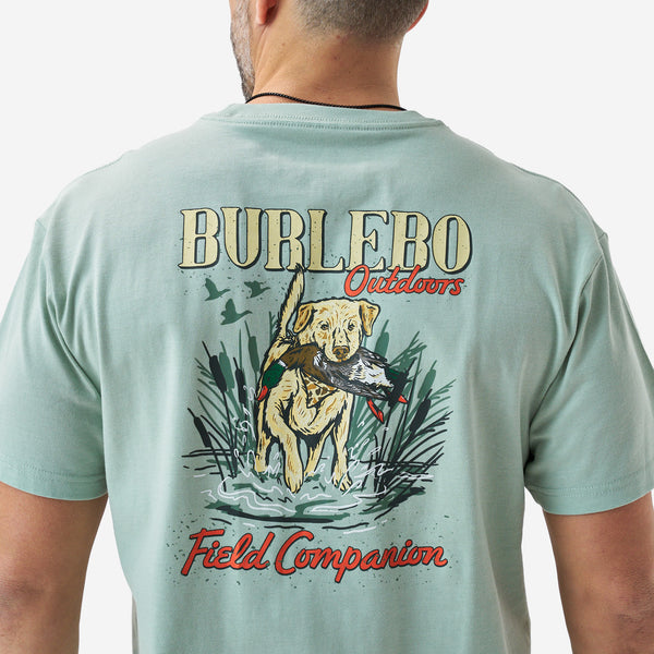 Back view of a male model wearing a sage green short sleeve Burlebo t-shirt featuring a “Burlebo Outdoors Field Companion” graphic with a retriever holding a duck in a marsh scene.