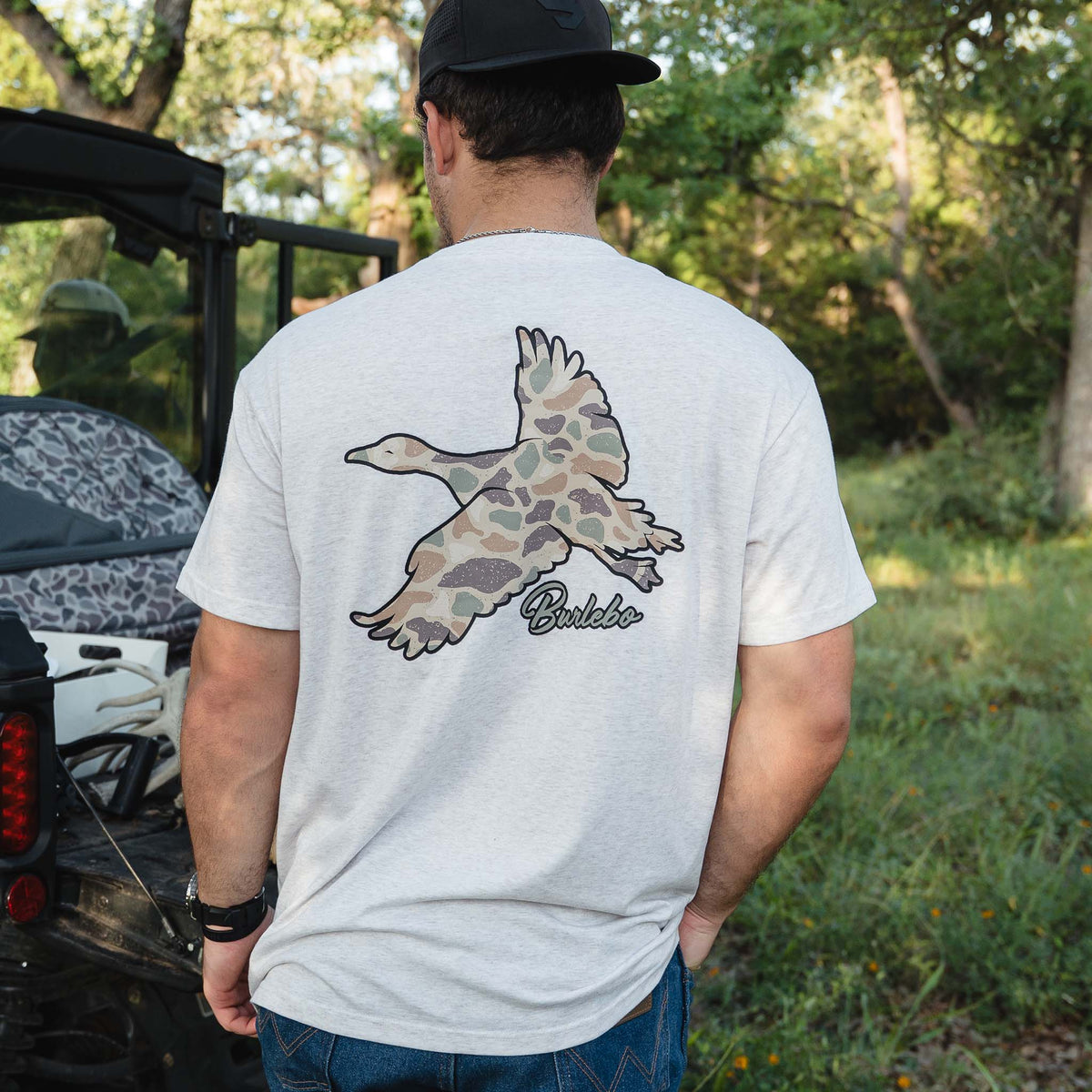 Light oat t-shirt with large camo duck graphic and "Burlebo" text on back, man standing outdoors near ATV.