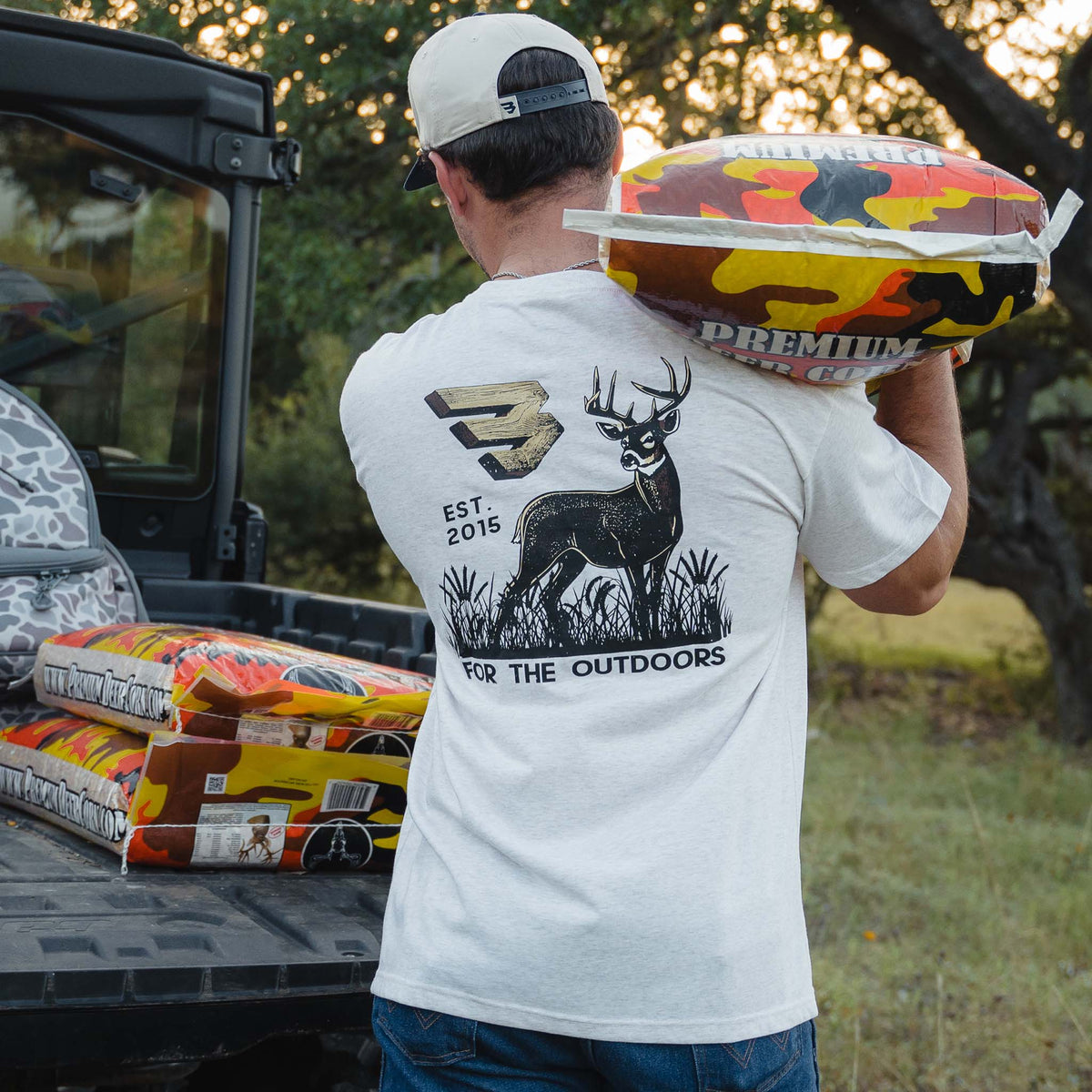 Light oat t-shirt with large back graphic of deer, camo "B" logo, "Est. 2015," and "For the Outdoors" text, man carrying camouflage feed bag over shoulder.