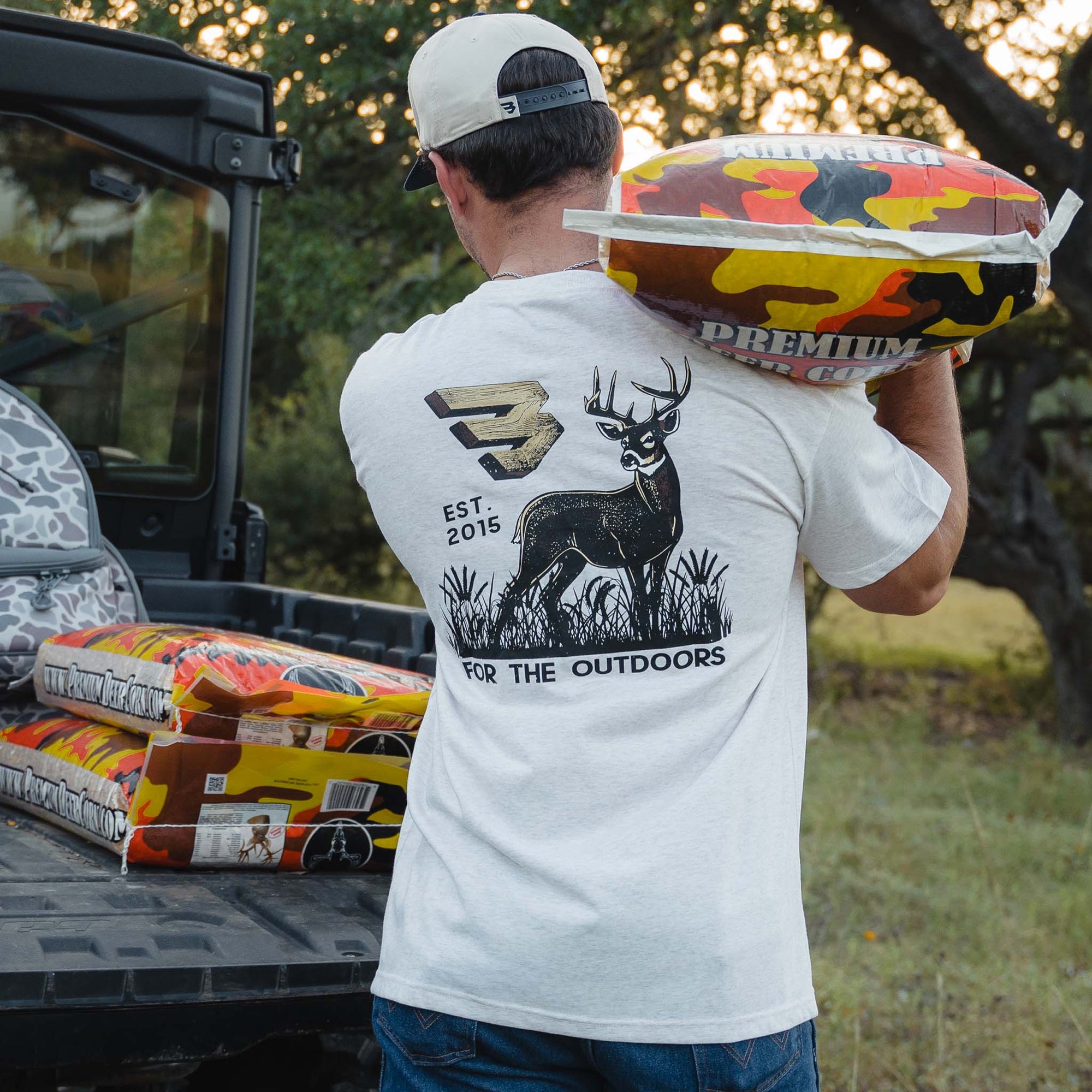 Light oat t-shirt with large back graphic of deer, camo "B" logo, "Est. 2015," and "For the Outdoors" text, man carrying camouflage feed bag over shoulder.