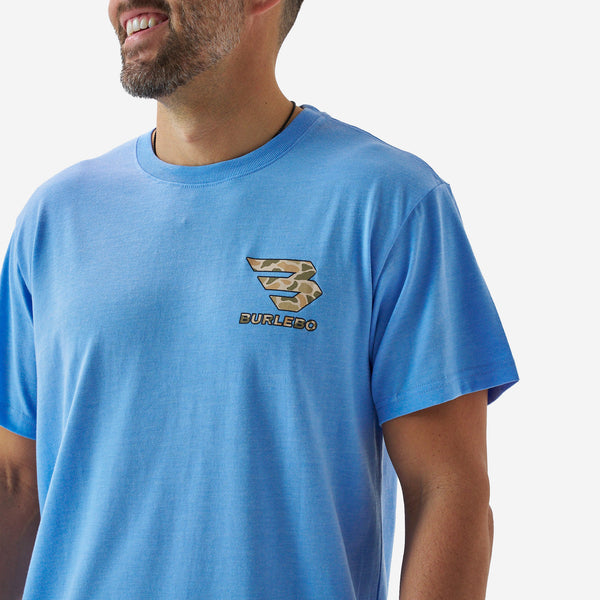 Close-up of a male model wearing a bright blue short sleeve Burlebo t-shirt with a small camouflage Burlebo logo printed on the chest.