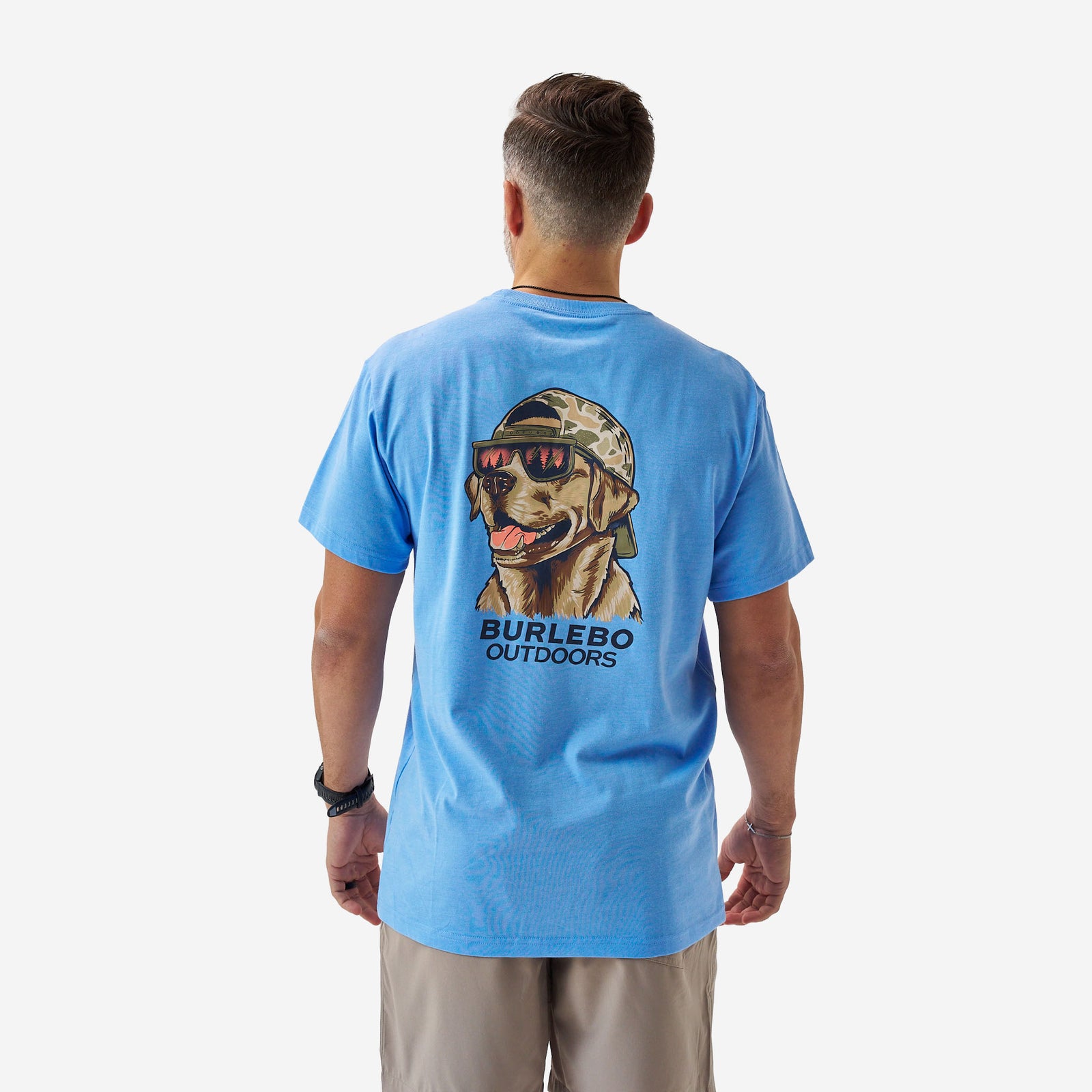 Rear full-body view of a male model wearing a bright blue short sleeve Burlebo t-shirt featuring a large graphic of a dog wearing sunglasses and a camo hat with “Burlebo Outdoors” text.